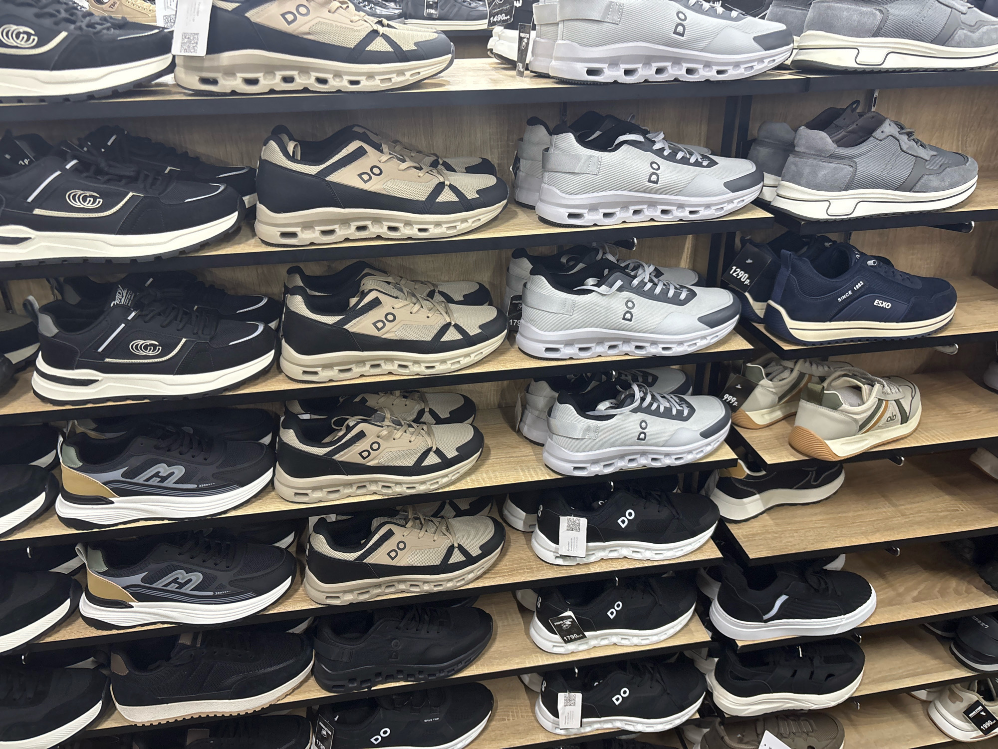 A multiple-shelf display of athletic shoes is visible in this indoor setting. The shelves are made of light-colored wood and are organized in a tiered fashion, with several rows of shoes on each level. The shoes are predominantly in shades of black, white, gray, and tan, with various designs and brand logos visible. Some shoes are positioned upright, while others are placed on their sides. The overall lighting suggests an indoor retail environment, likely a shoe store. The specific location in Mytishchi, Russia cannot be determined from the provided image.