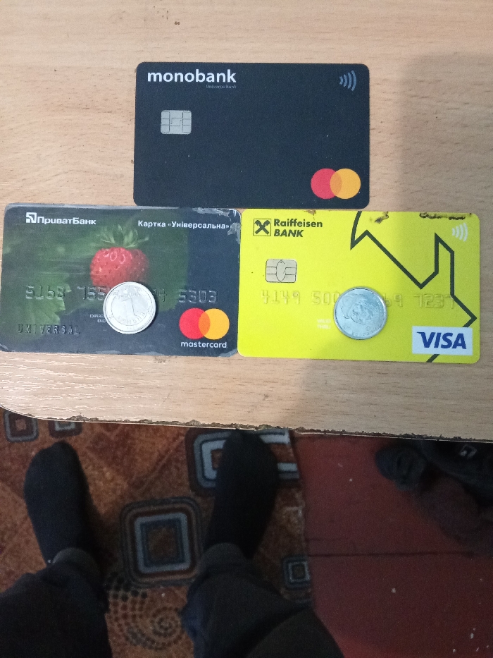 The image displays an indoor scene in the city of Yarun, Ukraine, featuring three Ukrainian bank cards and two coins arranged on a light-colored wooden surface. A black "monobank" card, horizontally oriented with a chip and contactless symbol, is positioned centrally at the top. Below it, to the left, is a dark grey "ПриватБанк" (PrivatBank) Mastercard depicting a large strawberry, showing a chip and partial numbers "5153-155" and "5303". To the right, a yellow "Raiffeisen BANK" Visa card, also horizontally oriented, shows a chip, contactless symbol, partial numbers "4149 500" and "123", and has minor damage along its top edge. A silver-colored Ukrainian 1 Hryvnia coin rests on the "ПриватБанк" card, partially obscuring the strawberry and numbers. Another silver-colored Ukrainian 1 Hryvnia coin rests on the yellow "Raiffeisen BANK" card, partially obscuring its numbers. The lower portion of the frame reveals a person's lower legs and feet, clad in black socks and dark pants, standing on a patterned rug with brown and geometric square designs. An adjacent reddish-brown floor area, possibly wood or linoleum, is visible to the right of the rug, with a dark shoe partially visible on it.