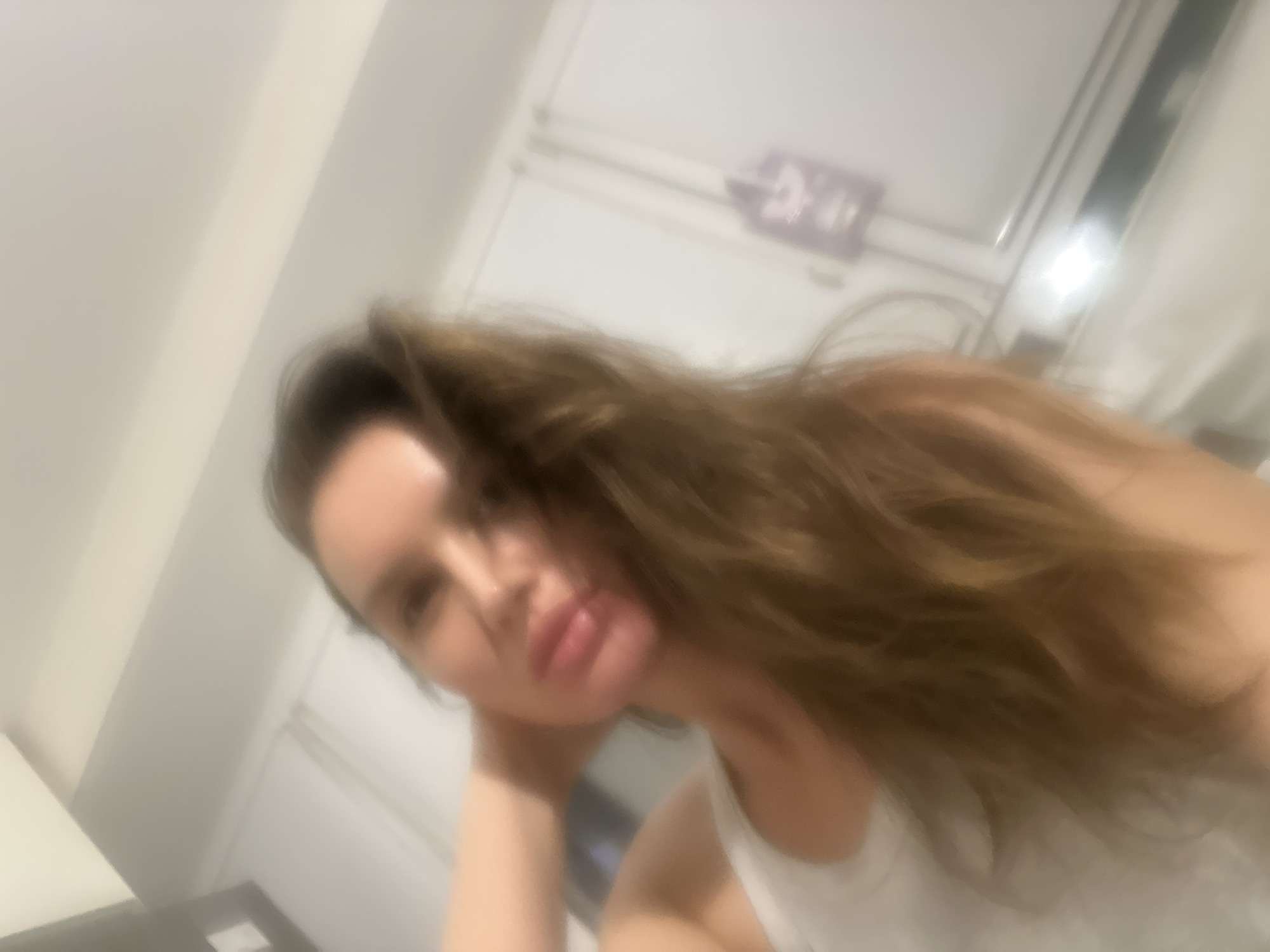 A highly blurry, low-focus indoor selfie captures a woman with long, wavy brown hair. The woman's face, with full lips, is angled towards the camera, and she appears to be resting her head on her hand or arm. Her expression is calm and relaxed, with a soft gaze. She is wearing a light-colored, possibly white, top or tank top.
The background is largely indistinct due to the blur, showing light-colored walls or a ceiling, indicating an indoor setting. In the upper right background, there are faint outlines of what could be a window or door frame. A dark, blurry shape in the lower left might be furniture, such as a table or dresser. On the wall in the upper background, there is a very faint, rectangular purple object that resembles a sign or picture, but any text or symbols on it are entirely illegible due to the lack of focus. The lighting is soft and diffused, typical of indoor artificial light, making the specific time of day unclear. The image depicts a casual, relaxed moment in an indoor environment, located in Moscow, Russia.