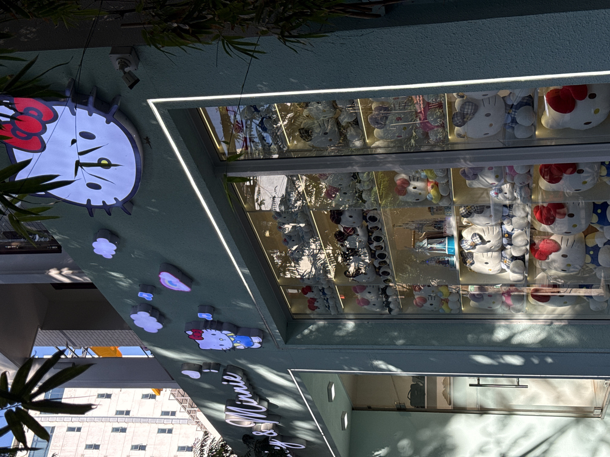 The image shows the exterior of a store in Đà Nẵng, Vietnam, identified by the signage "Hello & Minnie." The shop's façade features prominently displayed Hello Kitty merchandise, including plush toys visible through a window. A large Hello Kitty clock adorns the exterior, along with decorative cloud shapes. The scene includes foliage partially obscuring the view, suggesting an urban setting with vegetation. A building is visible in the background.