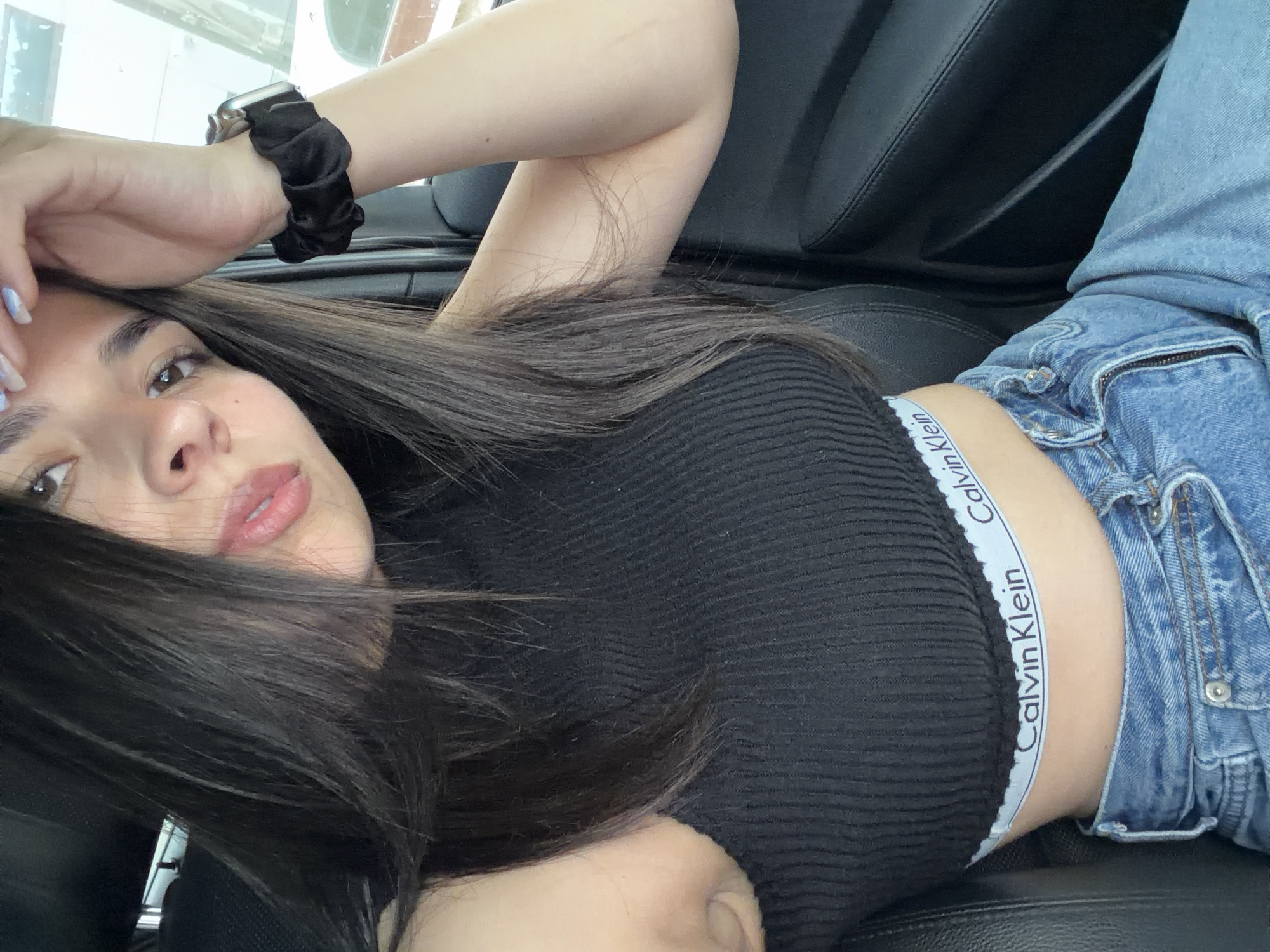 A young woman is captured in a close-up, slightly angled selfie, appearing to be lying down in a car. She has long, dark hair that partially covers her face and chest, light brown eyes, and her lips are slightly parted with a glossy finish. Her right hand is raised, resting gently on her forehead near her dark eyebrows. On her left wrist, a black scrunchie is worn along with a silver-cased smartwatch. Her fingernails are painted a light, iridescent blue.
She is dressed in a black, ribbed crop top that reveals her midriff, paired with light blue denim jeans. A notable detail is the elastic band of her undergarment or bralette peeking out above her jeans, clearly displaying the brand name "Calvin Klein" in white letters on a light-colored band.
The setting appears to be the interior of a vehicle, suggested by the black leather seats that form the background. The lighting indicates it is daytime, with some indistinct shapes and light visible through what looks like a car window in the upper left. The woman's expression is calm and direct, engaging with the camera. The location context for this image is Gabrovo, Bulgaria.