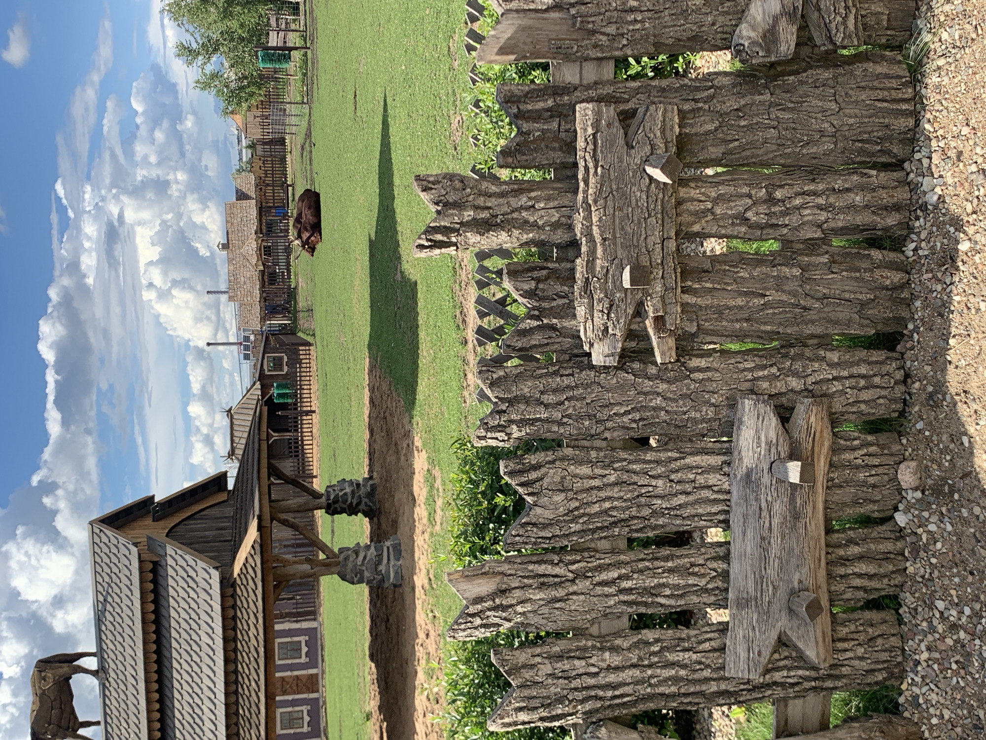 The image depicts an outdoor scene at a Belarusian park or attraction, likely in or near Малахавецкі сельскі Савет. In the foreground, a rustic fence constructed from rough-hewn logs with prominent bark texture runs horizontally across the frame. Attached to the fence are two wooden decorative elements, one resembling a stylized animal head. Behind the fence, a lush green lawn stretches into the middle ground, where a few dark brown animals, possibly bison or cows, are lying down. Further back, a cluster of rustic wooden buildings with shingled roofs and smaller fences suggests a village or historical settlement setting. In the background, a blue sky with scattered white clouds provides a backdrop, with hints of industrial structures and trees on the horizon. The overall impression is of a well-maintained rural or historical park with animal enclosures and themed architecture.