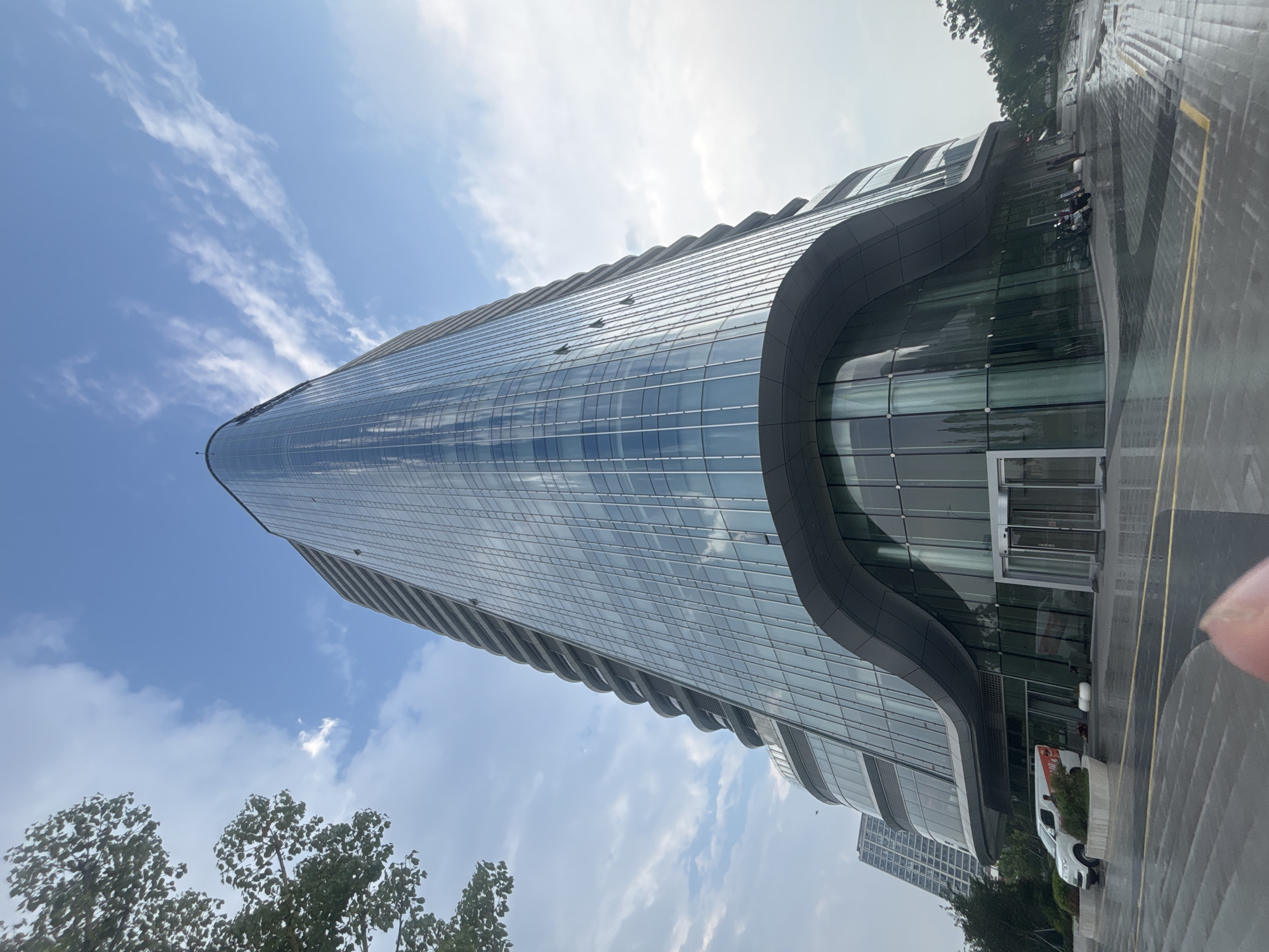 The image shows a modern, curved glass building in Suzhou Industrial Park, China, on a cloudy day. The building's facade is made of reflective glass, creating a striking geometric pattern. A white van is parked in front of the building, and several motorcycles are visible near the entrance. The ground is wet, suggesting recent rain. Trees and other buildings can be seen in the background, indicating an urban environment.