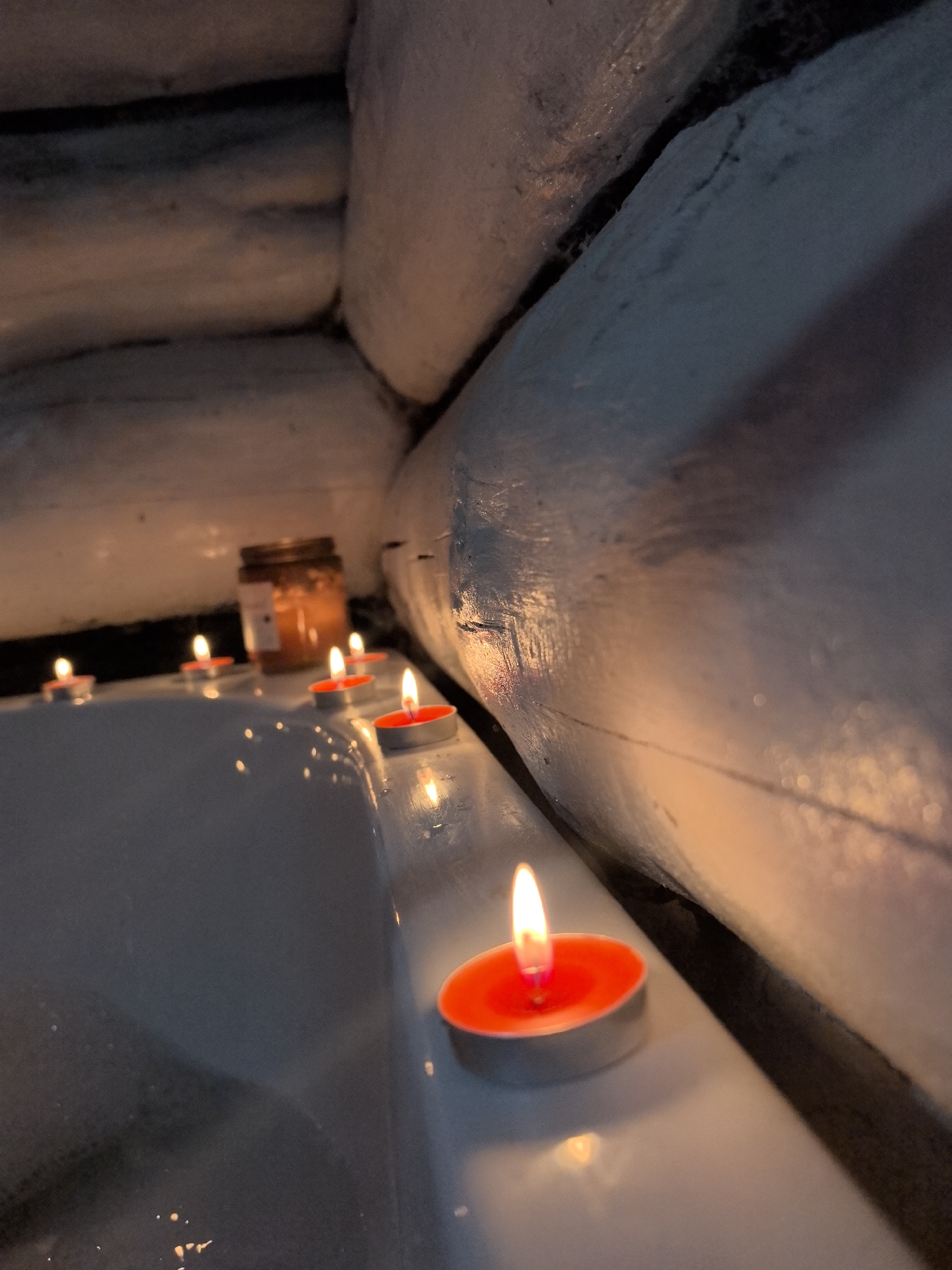 A close-up shot depicts a bathtub rim illuminated by several small, lit candles. The candles have red wax and are encased in silver aluminum containers. A glass jar sits among the candles, containing a brownish substance. The bathtub is white and appears to have some water or condensation on its surface. The surrounding environment suggests a rustic or cabin-like setting, with white-painted logs forming the walls. The overall ambiance is warm and intimate, created by the soft glow of the candles. There is no evidence to suggest the image was taken in Noviy, Russia.