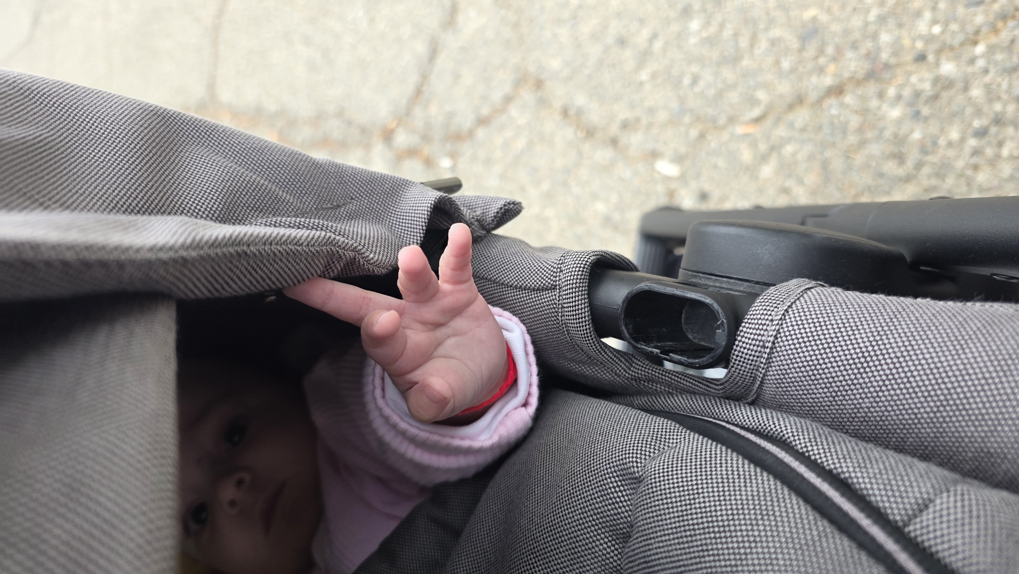 A close-up shot shows a baby's hand reaching out from under the canopy of a gray stroller. The baby's face is partially visible, indicating they are lying inside the stroller. The stroller's frame and canopy are made of gray patterned fabric and black plastic components. The background is blurred, showing a paved surface. It is not possible to determine the location in the city of Blagoevgrad, Bulgaria from the image.