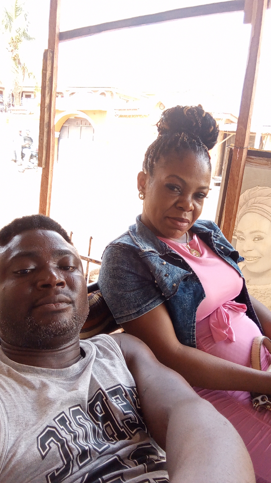 Two people are captured in a selfie. A man in a gray tank top is in the foreground. A woman wearing a pink dress and a denim jacket is seated behind him. Her hair is styled in a high bun. An framed image of a woman's portrait is behind her. They appear to be sitting in an outdoor setting with a partially visible building and trees in the background. This image was taken in Sabon Tasha, Nigeria.