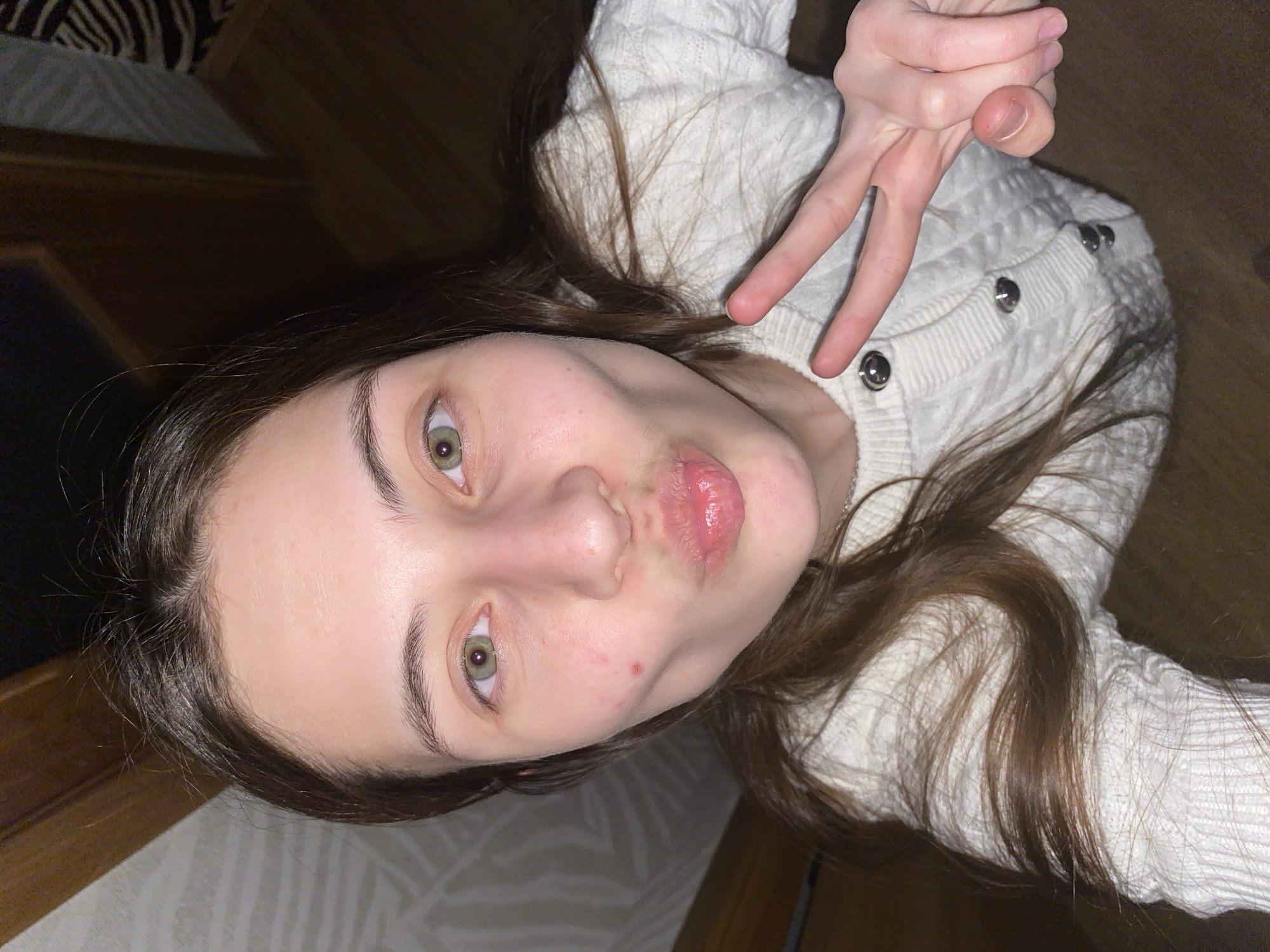 A young woman is captured in a close-up, angled selfie, likely taken from above while she is lying down. She has fair skin, long brown hair, and striking green eyes, looking directly at the camera. Her lips are puckered in a playful expression, and she is making a peace sign gesture with her right hand, which is visible in the upper right quadrant of the image.
She is wearing a white, possibly cream-colored, cable-knit sweater or cardigan, which features silver buttons along the shoulder area. The background suggests an indoor setting, likely a home, with dark wooden elements visible on one side and a patterned surface, possibly a wall or bedding, on the other. The lighting is soft and ambient, indicating an interior shot. There is no visible text in the image. The overall mood conveyed is casual and relaxed, with a touch of playfulness. The location context provided is Votkinsk, Russia.