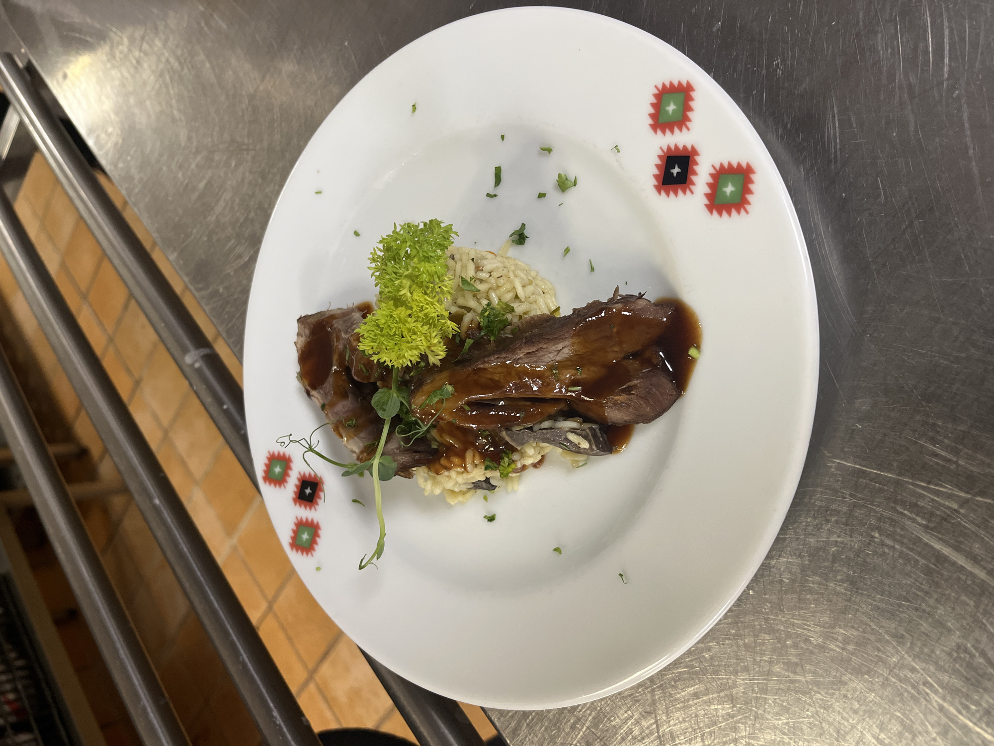 A close-up, eye-level shot shows a white plate of food placed on a brushed metal surface. The plate is adorned with three small, square, decorative patterns along its rim, featuring red and green zig-zag borders around a central black or green square with a white cross.
The food on the plate consists of what appears to be sliced roast meat covered in a dark, glossy sauce, served atop a bed of rice pilaf. A small sprig of bright green curly parsley or similar herb garnishes the dish, along with a few delicate green sprouts and finely chopped green herbs scattered across the plate. The meat is dark brown and appears tender, with the sauce pooling around it. The rice is a pale yellow-white, with visible grains and what seem to be small pieces of mushroom or other dark ingredients mixed in.
The background of the image is out of focus, but shows what appears to be a commercial kitchen environment. Two horizontal metal bars are visible above and behind the plate, and beyond those, a tiled wall with a warm, orangey-brown hue. The brushed metal surface beneath the plate is scratched and shows signs of wear. The lighting is bright, highlighting the food and creating reflections on the metal surface. There are no people visible in the image.