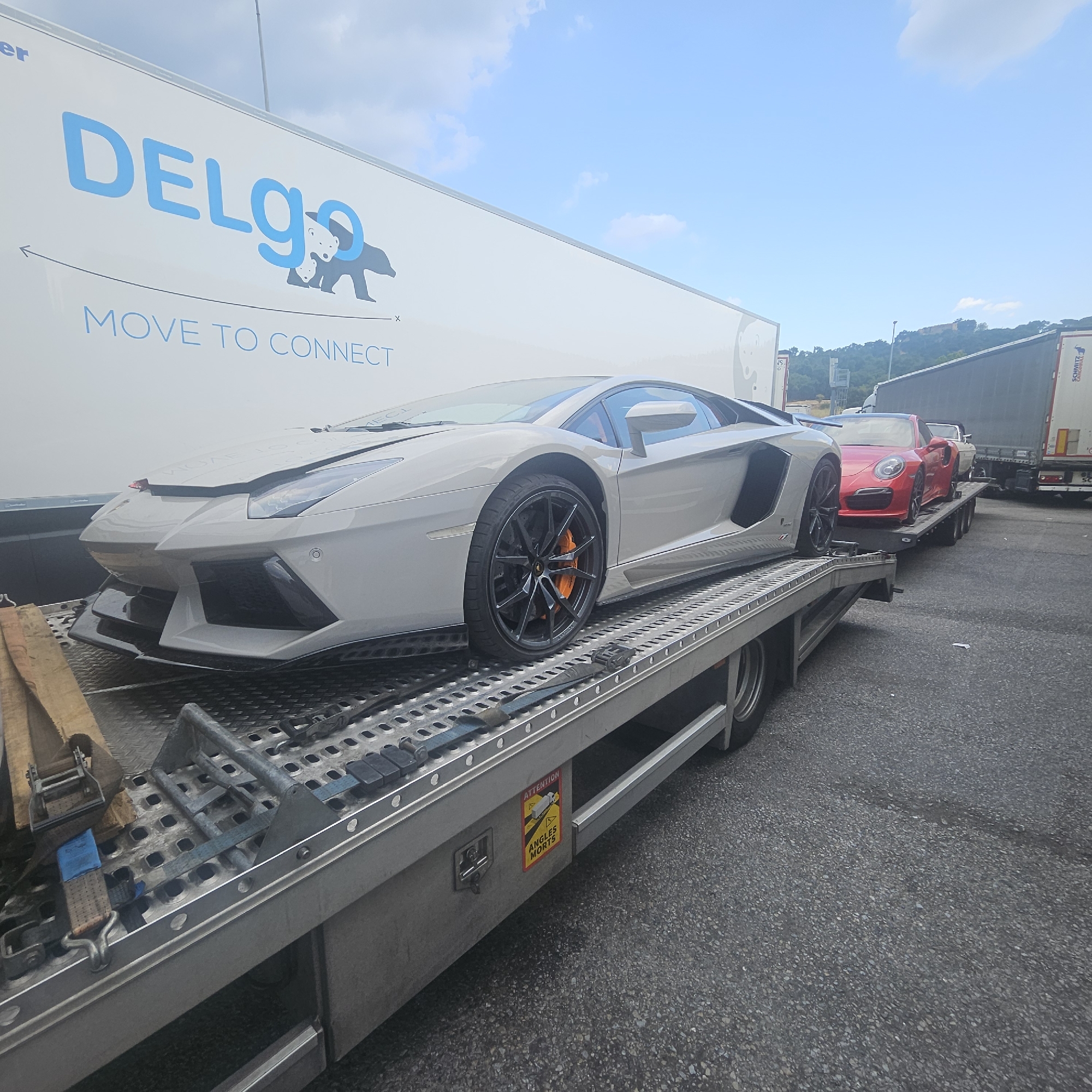 In La Jonquera, Spain, a light gray Lamborghini Aventador is loaded onto a car carrier trailer. The trailer is parked on an asphalt surface under a partly cloudy sky. The Lamborghini is the primary subject, positioned in the foreground, with its front fascia, headlights, and a portion of its side visible. The car is secured to the trailer with straps.

Behind the Lamborghini, a red Porsche is also loaded onto a trailer. The Porsche is positioned further back and is largely visible. Another vehicle, possibly a white convertible, is behind the Porsche, partially obscured.

The trailer itself is silver and metallic, with a checkered plate surface designed for vehicle transport. Straps and tie-down mechanisms are visible securing the vehicles. To the left of the Lamborghini, a large white trailer with blue lettering that reads "DELGO MOVE TO CONNECT" is visible. The trailer has a logo featuring two bears.

A yellow and red warning sticker with the text "ATTENTION ANGLES MORTS" (French for "Attention Blind Spots") is affixed to the side of the car carrier trailer. In the background, the scene includes other trucks, one with "SCHMITZ" branding, and a hillside covered in green foliage. The overall impression is a commercial transport operation.