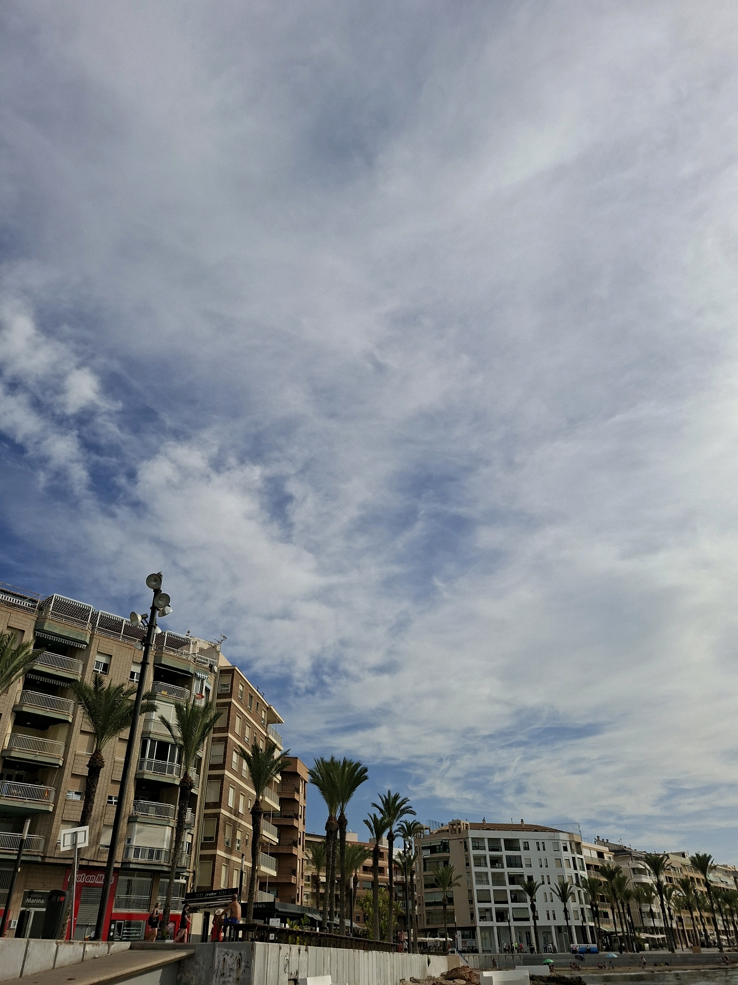 The image captures a street scene in Torrevieja, Spain, characterized by a row of multi-story buildings and palm trees lining what appears to be a promenade or waterfront. The buildings are predominantly made of brick and feature balconies and large windows, indicative of residential or commercial spaces. Several tall palm trees are scattered throughout the scene, adding a tropical element. In the foreground, there is a concrete barrier or wall, suggesting a separation from a lower level or the sea. A few people are visible in the middle ground, some walking and others seated, near what looks like outdoor seating for a cafe or restaurant. The sky above is a mix of blue and white clouds, suggesting fair weather. The overall impression is a bustling urban environment near the coast, common in Mediterranean coastal towns.