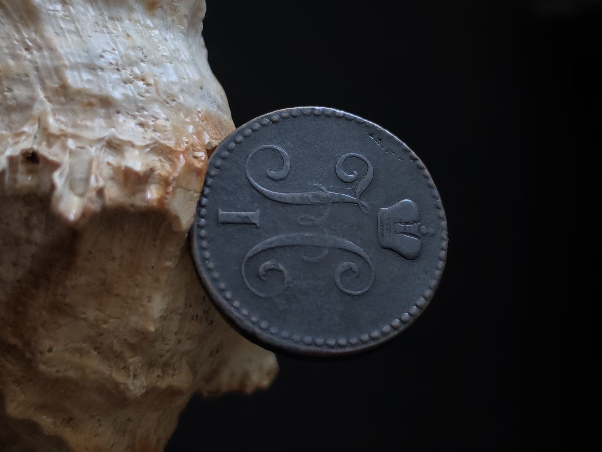 A close-up, dimly lit photograph shows the obverse of a dark, circular coin resting on a light-colored, textured surface that resembles a seashell or coral fragment. The coin is centrally positioned and angled, revealing a crowned monogram composed of intertwined letters and the numeral "1" below it. The background is uniformly dark and out of focus, suggesting a controlled studio environment. The lighting highlights the metallic sheen and intricate details of the coin. The overall impression is one of historical artifact presentation. No specific location in Strelna, Russia, can be determined from the visual information provided.
