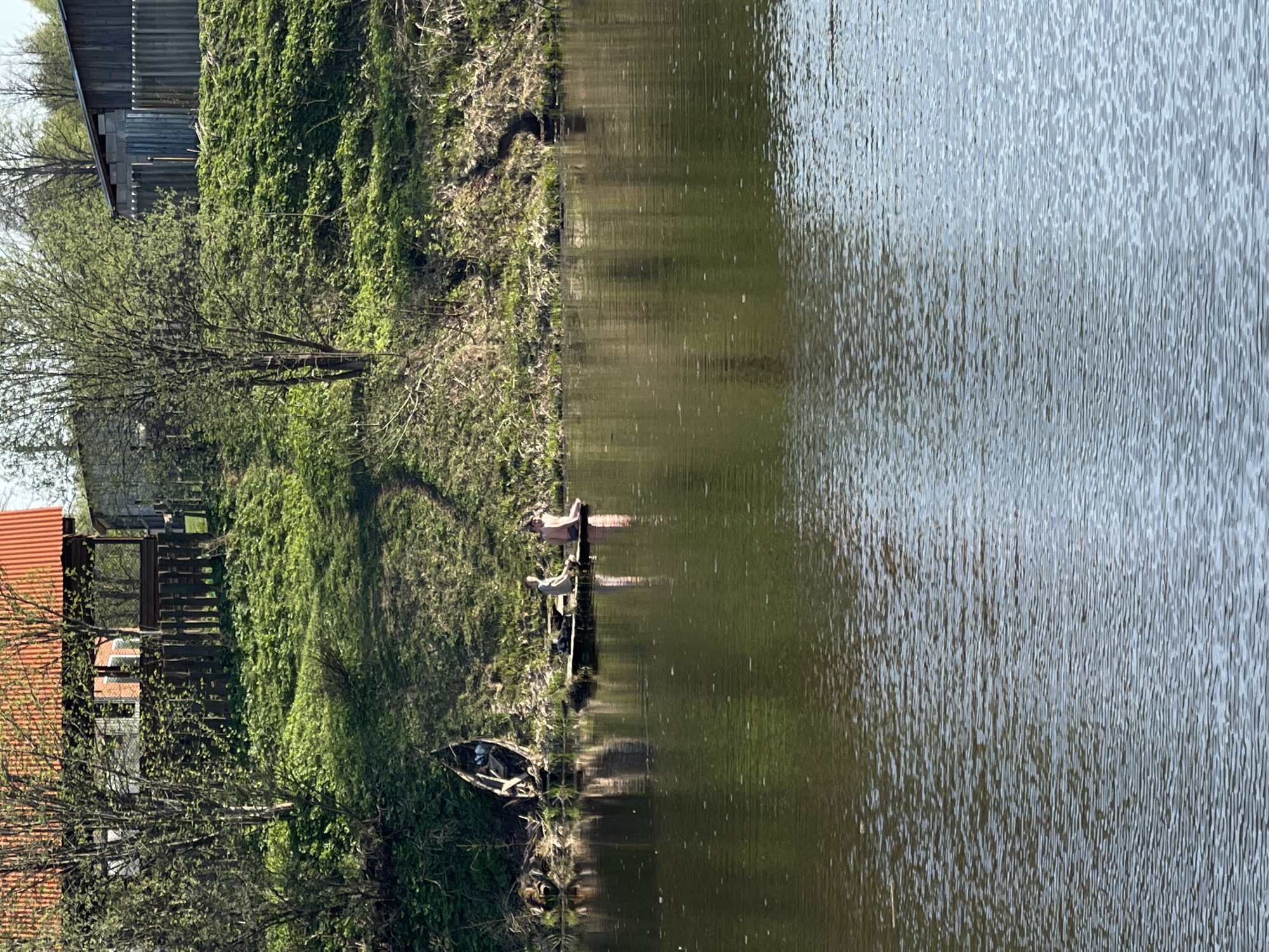 The image shows a tranquil scene at Zhigalki, Russia. A body of water, possibly a lake or wide river, dominates the right side of the frame, its surface reflecting light and surrounding greenery. The left side depicts a grassy bank leading up to a wooded area. A small wooden dock extends from the bank into the water. Two people are present on the dock, one appearing to be lying down, while the other is sitting. Closer to the bank, an old, damaged boat rests along the shoreline. In the background, structures resembling a house with a red roof and other buildings behind a fence are visible. The overall impression is one of a quiet, rural setting.