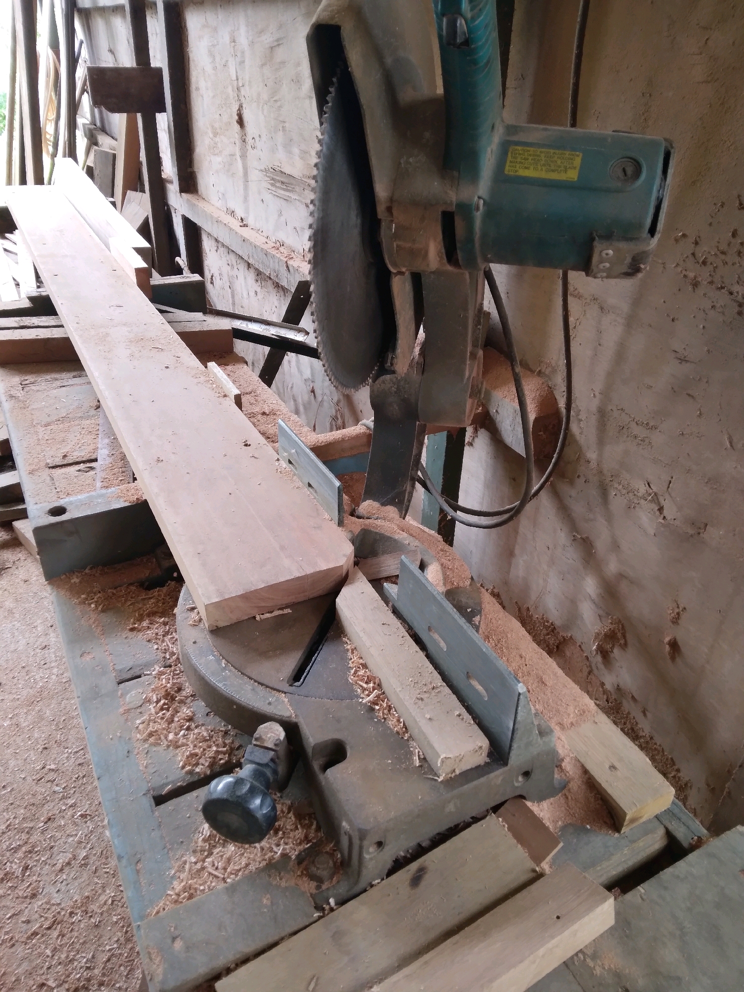 The image depicts a miter saw setup in what appears to be a workshop or construction site, likely in Batam, Indonesia. A long piece of light-colored wood rests on the saw's table, positioned for cutting. Several smaller pieces of wood are also present, some on the saw and others nearby. Sawdust is scattered across the saw's surface and the surrounding area, indicating recent activity. The background consists of a wooden wall and structural elements, suggesting an enclosed or semi-enclosed workspace. The lighting is natural, possibly from an open side of the structure, casting some shadows. There are no people visible in the image. The overall scene conveys a utilitarian and working environment focused on woodworking.