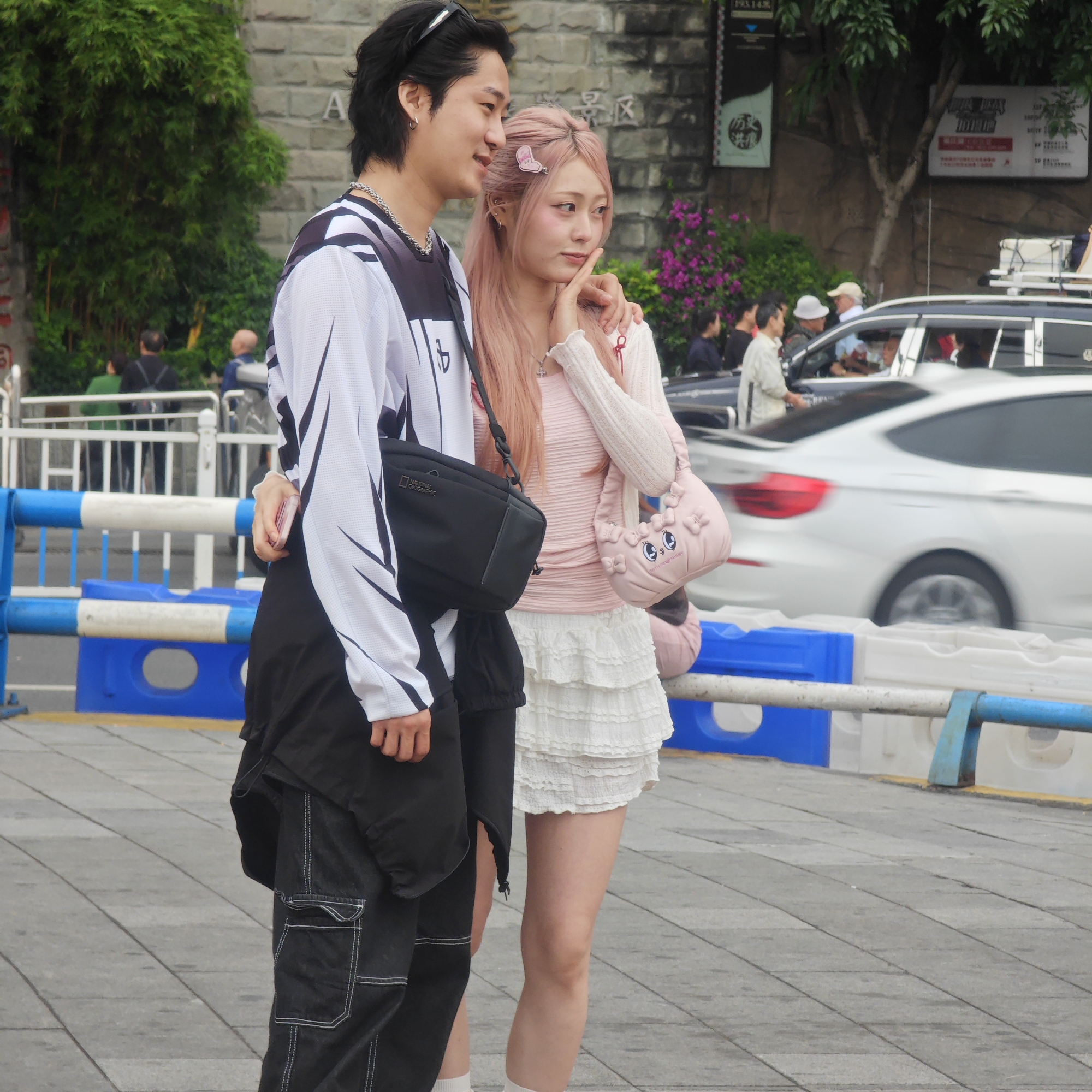 Observed at Yuzhong District, China, are two individuals standing on a grey tiled ground next to a blue and white barrier. The person on the left is male with black hair and wears a white long-sleeved top with black patterns, a black jacket tied around the waist, and black pants with white stitching. A black shoulder bag is slung across his body. His arm is around the shoulders of the person to his right. The person on the right is female with pink hair and a pink hair clip. She wears a pink top, a white cardigan, and a tiered white skirt. She carries a pink purse with a cartoon face. In the background, there are other people, vehicles, trees, and a stone wall with signs.