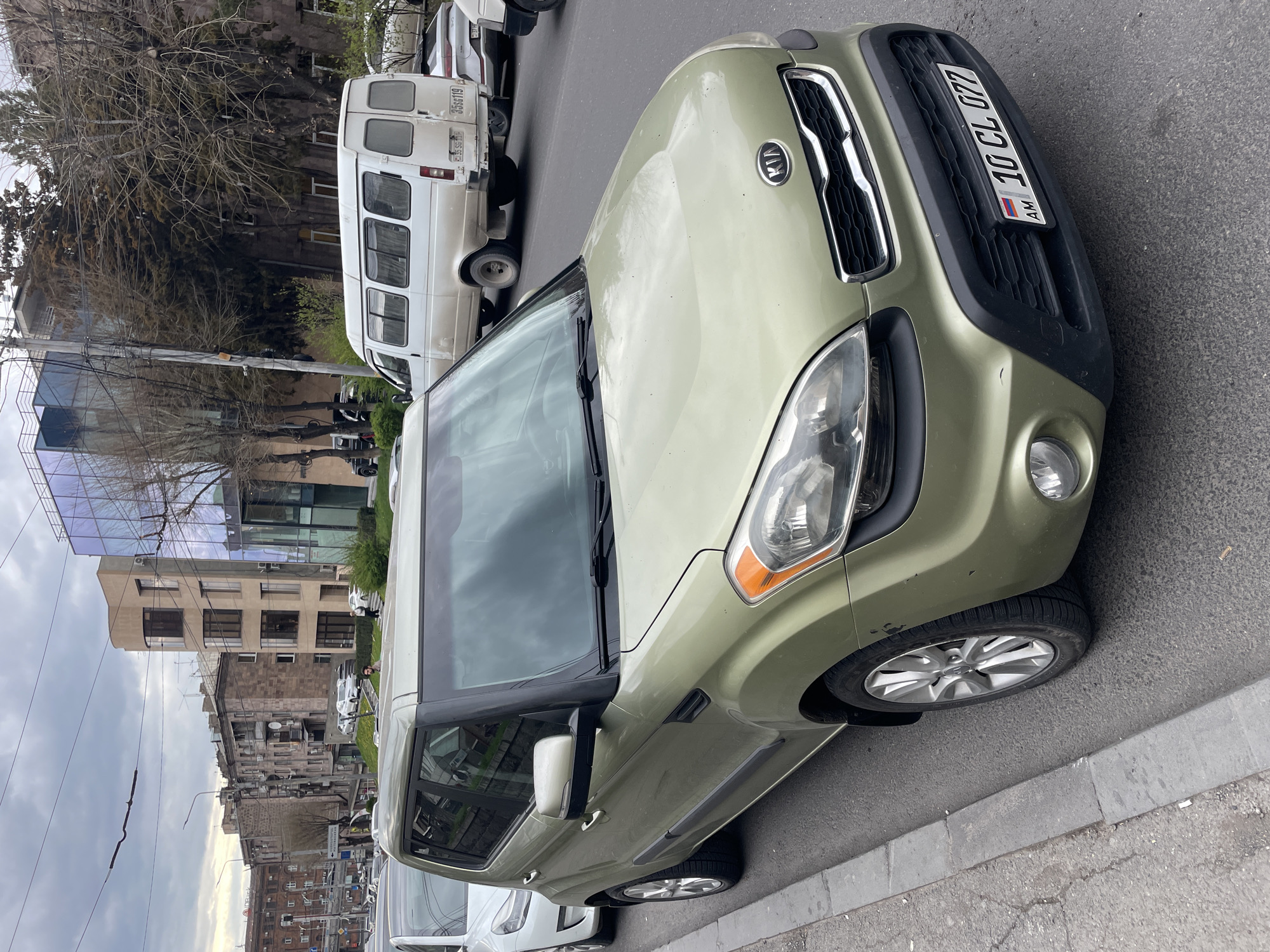A light-green Kia Soul is parked on a city street in Yerevan, Armenia. Its license plate reads "AM 10 CL 077". A white van is parked behind it. The scene is an urban environment, showing buildings of varying architectural styles and ages, parked vehicles, and a cloudy sky.