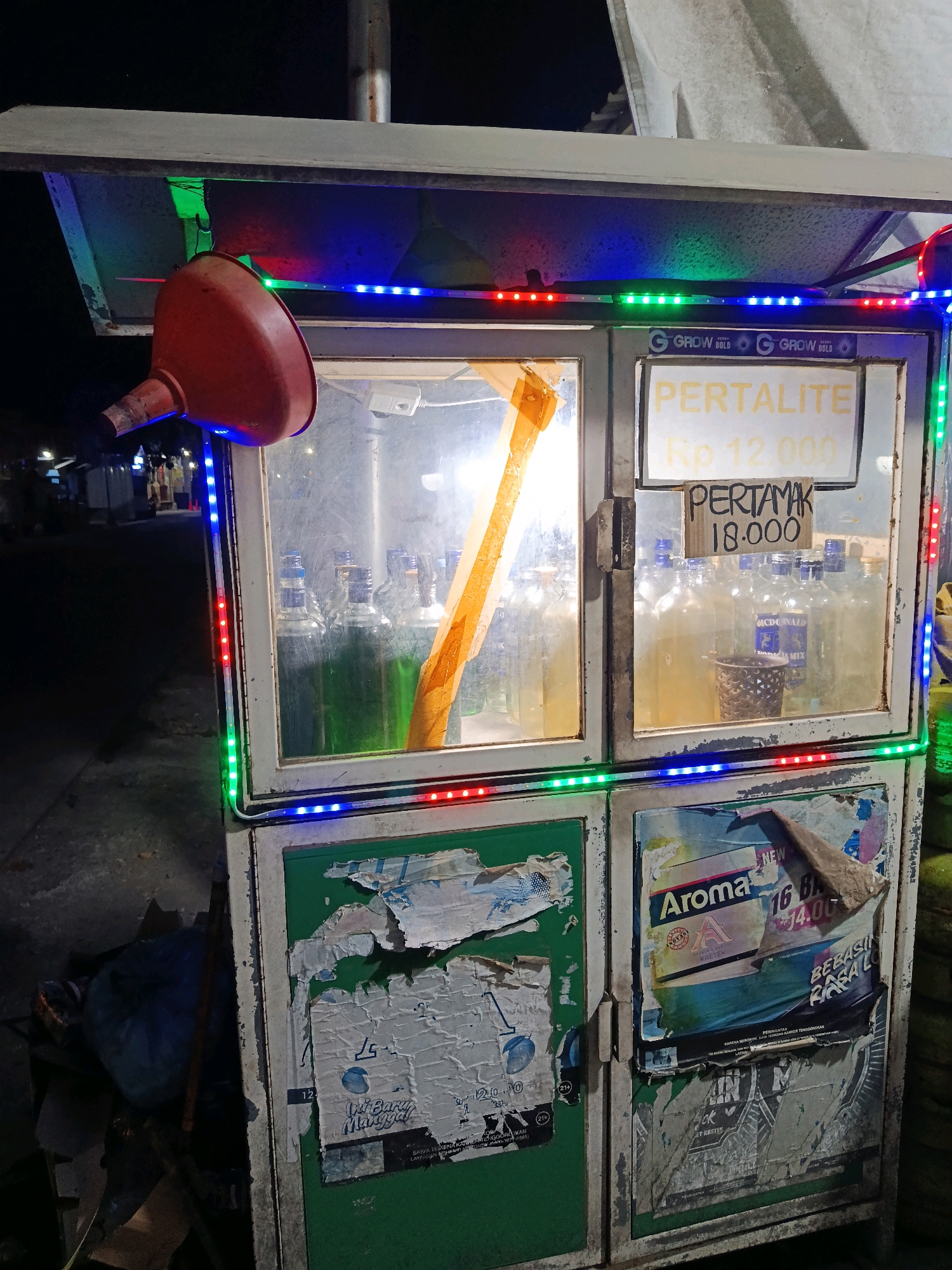 An outdoor night scene depicts a portable fuel stand for sale in Demak, Indonesia. The stand is illuminated by multicolored LED lights around its upper edge. A red funnel hangs from the top left of the stand. The stand has a glass display case showcasing numerous clear and green bottles filled with liquid. Signage on the front of the stand indicates "PERTALITE Rp 12.000" and "PERTAMAK 18.000". Below the display case are two cabinets, the left one being green and showing torn posters, and the right one also displaying torn posters, including one with the word "Aroma" prominently featured. The ground around the stand is paved, and a blue plastic bag and other debris are visible to the left. In the background, a dimly lit street with buildings and trees suggests a residential or commercial area.