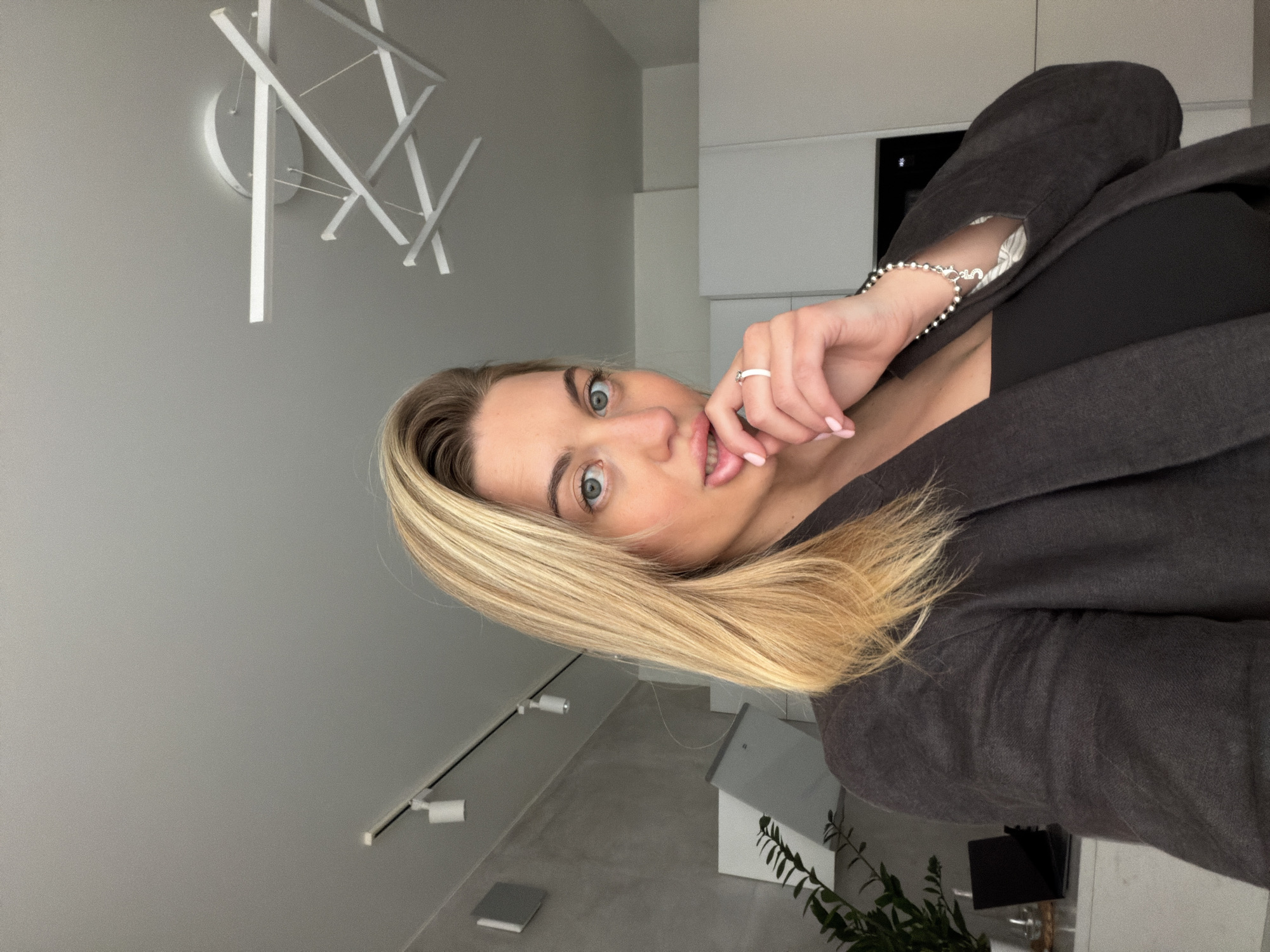 The image captures a close-up of a woman with blonde hair and striking blue-green eyes, looking directly at the camera with an expressive, slightly surprised or playful gaze. Her head is tilted, and her right hand is brought up to her face, with manicured, light pink nails touching her lower lip. She wears a silver-toned ring with a white detail on her ring finger and a beaded silver bracelet with a decorative charm on her wrist. Her attire includes a dark top and a dark grey or charcoal blazer.
The setting is a modern, minimalist interior, likely an apartment or house located in Sofiivska Borshchahivka, Ukraine. The walls are a light grey. In the background, to the upper left, a contemporary ceiling light fixture with geometric white linear elements is prominent. On the right side, sleek, white kitchen cabinets or built-in storage units are visible. The lower portion of the image reveals a light-colored floor, possibly concrete, and track lighting with two spotlights. Decorative elements include a small, white house-shaped object, a green plant, and a dark rectangular item on a surface. The lighting is bright and even, indicating an indoor daytime scene. No visible text is present in the image.