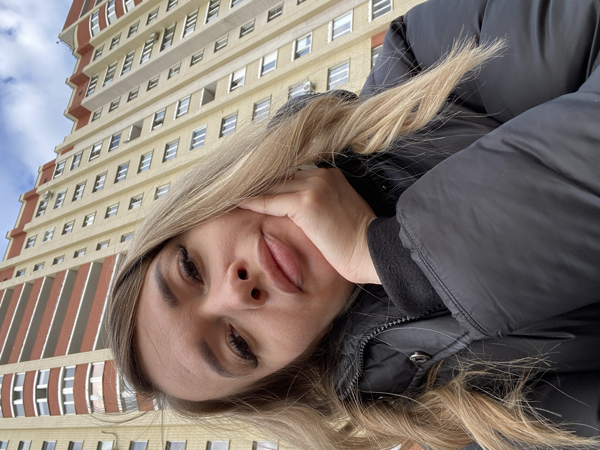 A person is holding their hand to their face while wearing a black puffer jacket. In the background, there is a tall building with many windows. The location is Izhevsk, Russia.