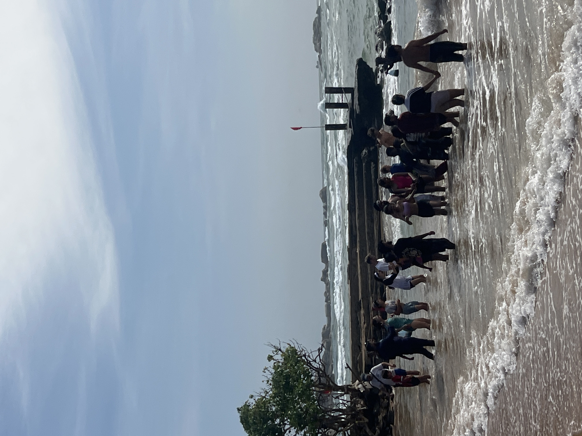 A group of approximately 20 people, varying in age, are standing in shallow, wavy ocean water near a rocky shore in Hikkaduwa, Sri Lanka. The people are dressed in casual beach attire. Some are walking, some appear to be standing and observing, and one pair in the foreground is holding hands. To the left, a lush green tree with bare branches stands near the water's edge. In the background, large rocks are visible in the ocean, and waves are crashing against a man-made structure with wooden posts and a red flag. The sky above is mostly clear with scattered white clouds. The scene depicts a casual gathering at a beach with moderately rough water conditions.