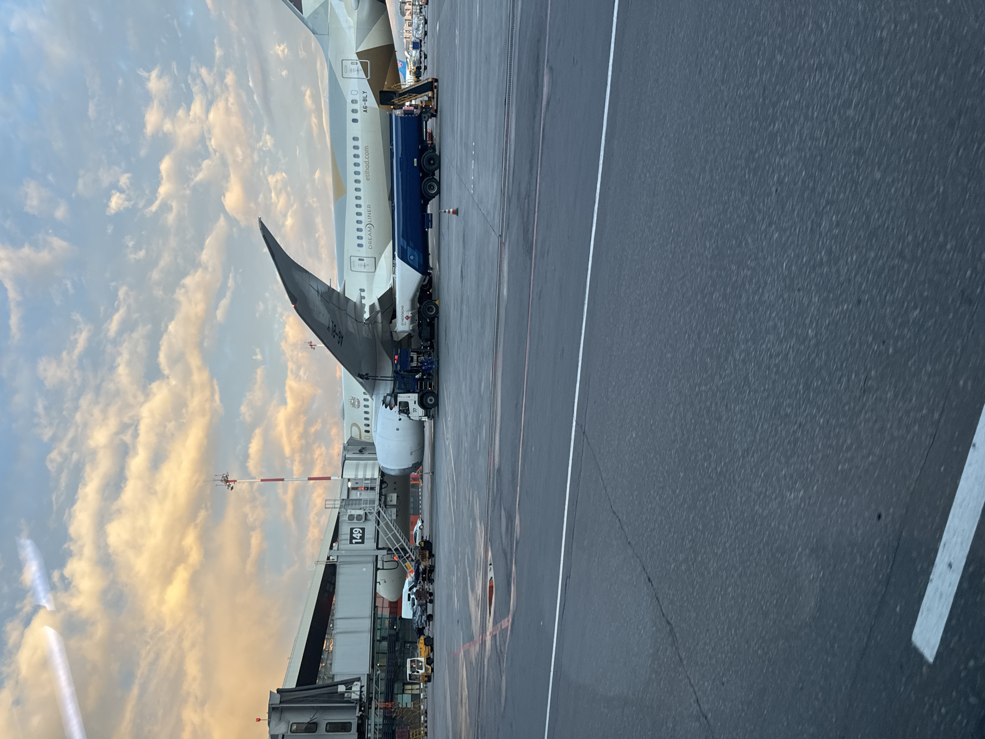 An Etihad Airways Boeing 787 Dreamliner, registration number A6-BLY, is parked at gate 149 at an airport. A large blue fuel truck is positioned next to the aircraft, possibly refueling it. Other airport vehicles are parked nearby. The sky is filled with clouds illuminated by the setting sun. The image was taken at an airport in Khimki, Russia.