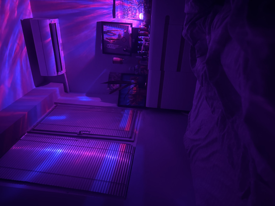 The scene depicts an interior room, likely a bedroom, in Novi Pazar, Bulgaria, illuminated predominantly by artificial blue and purple light, creating swirling patterns on the ceiling and walls. A window fitted with horizontal white blinds occupies the left side of the frame, reflecting blue and magenta light strips. An air conditioning unit is mounted high on the wall in the upper right quadrant. Below it, a white chest of drawers or dresser with multiple compartments is positioned against the wall. The dresser surface holds various objects, including two framed items (one with a potted plant in front of it), multiple bottles, cans, and a small illuminated object towards the right. The foreground and lower portion of the image reveal a rumpled bed with light-colored sheets, heavily saturated by the purple illumination. No individuals are present in the frame, and no discernible actions or interactions are occurring. The environment is orderly despite the scattered items on the dresser, suggesting a lived-in space under specific atmospheric lighting conditions.