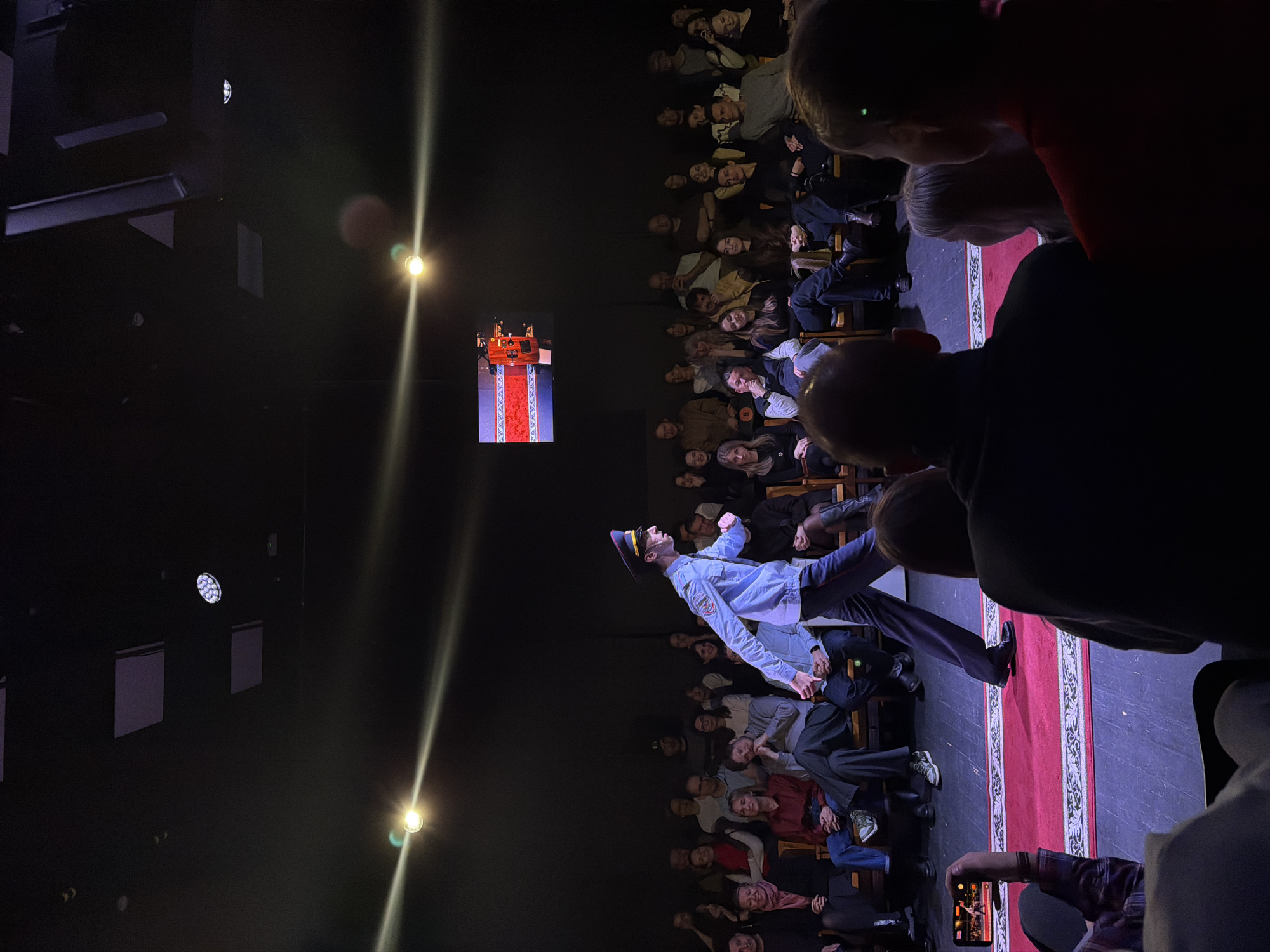 The image depicts an indoor theatrical performance captured from an audience member's perspective.
**Subjects:**
The main subjects are a male performer on stage and numerous audience members. The performer is dressed in a light blue uniform with a peaked cap, dark trousers, and black shoes, and appears to be in motion, leaning at a dynamic angle with one arm raised. The audience, consisting mostly of adults of varying ages, is seated in rows of wooden chairs, facing the stage. Many faces are visible, illuminated by the stage lights. In the immediate foreground, a person's head and shoulders are visible, and their hand holds a smartphone, apparently recording the event.
**Objects:**
The stage features a prominent red carpet with intricate white and gold patterns. Towards the rear of the stage, a small, ornate structure, resembling a throne or podium, is visible in red and gold. The background is dark, but various lighting fixtures are discernible: bright spotlights creating lens flare, and several circular and rectangular lights mounted on the wall or ceiling. The audience is seated on simple wooden chairs.
**Scene and Setting:**
The scene is set in a dark theater or performance hall. The artificial lighting, primarily focused on the stage, creates a dramatic atmosphere. The setting is intimate, with the audience relatively close to the stage.
**Activity or Event:**
A live performance is taking place on stage. The performer's pose suggests a dramatic or comedic act. The audience is actively watching and engaged, with at least one person recording the event on a mobile phone.
**Notable Details:**
The event appears to be occurring in the evening or at night, given the dark surroundings and reliance on artificial lighting. The audience members seem attentive, with some showing expressions of amusement or concentration. There are no specific textual elements clearly visible within the image, such as signs or posters. The context provided indicates the location is Moscow, Russia.