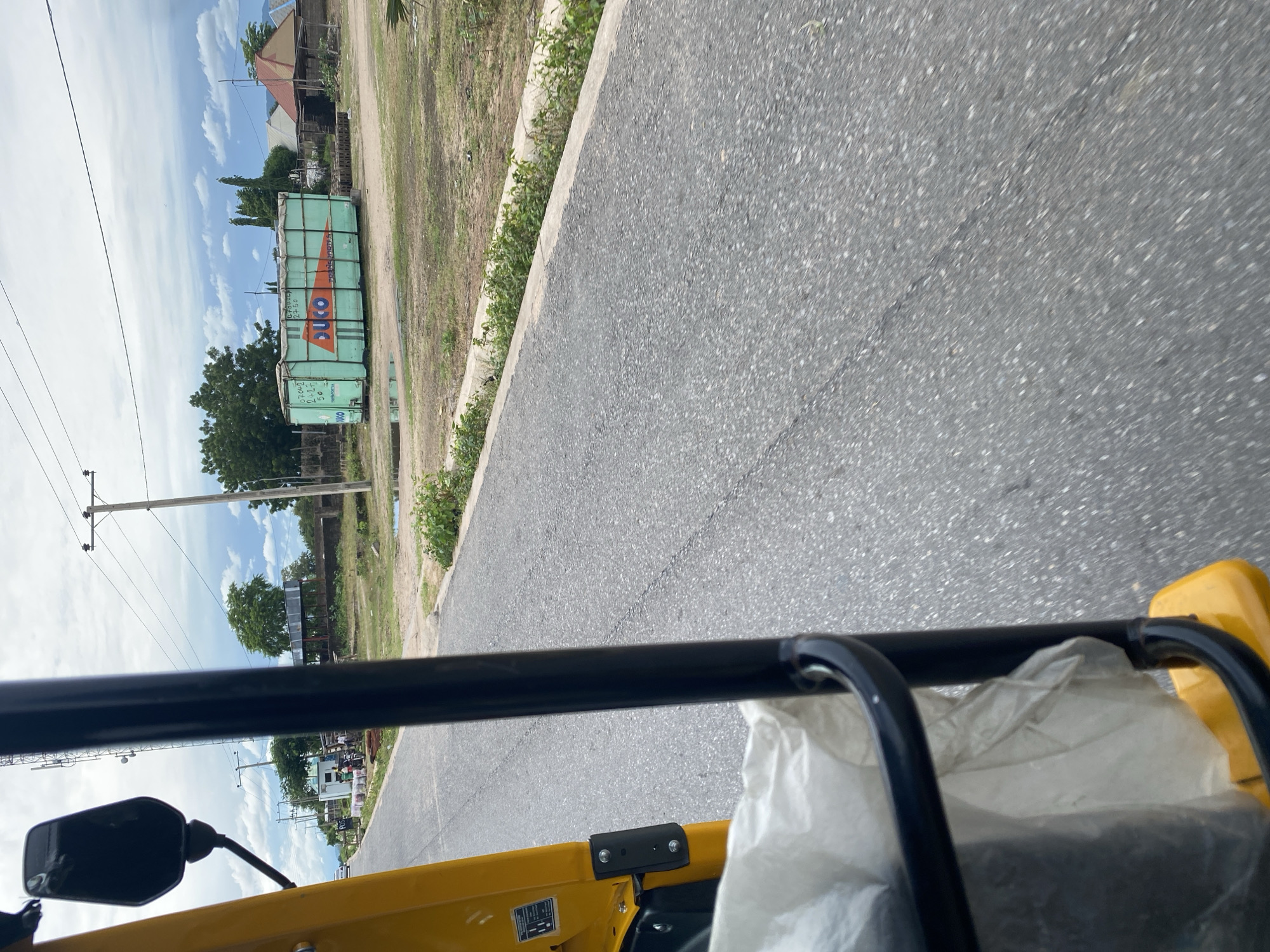 This media file is a photograph taken from the perspective of someone riding in a yellow auto-rickshaw. The scene is a road in Jalingo, Nigeria, with a grey asphalt surface in the foreground. To the left of the road, there is a dry, grassy area bordered by a concrete curb and some green vegetation. In the middle ground, several green shipping containers are stacked, one with the word "DUCO" prominently displayed in orange and white lettering. Above the containers, a blue sky with white clouds is visible. There are trees and some buildings in the background. Utility poles and power lines are also present along the roadside. The image appears to have been taken during daylight, likely in the morning or afternoon, given the brightness of the sky and the shadows. The weather seems to be clear and sunny. The activity taking place is travel, with the auto-rickshaw moving along the road. There are no people clearly visible in the image, though the presence of the auto-rickshaw suggests human activity. The overall impression is of a somewhat rural or semi-urban environment on a typical day in Jalingo.