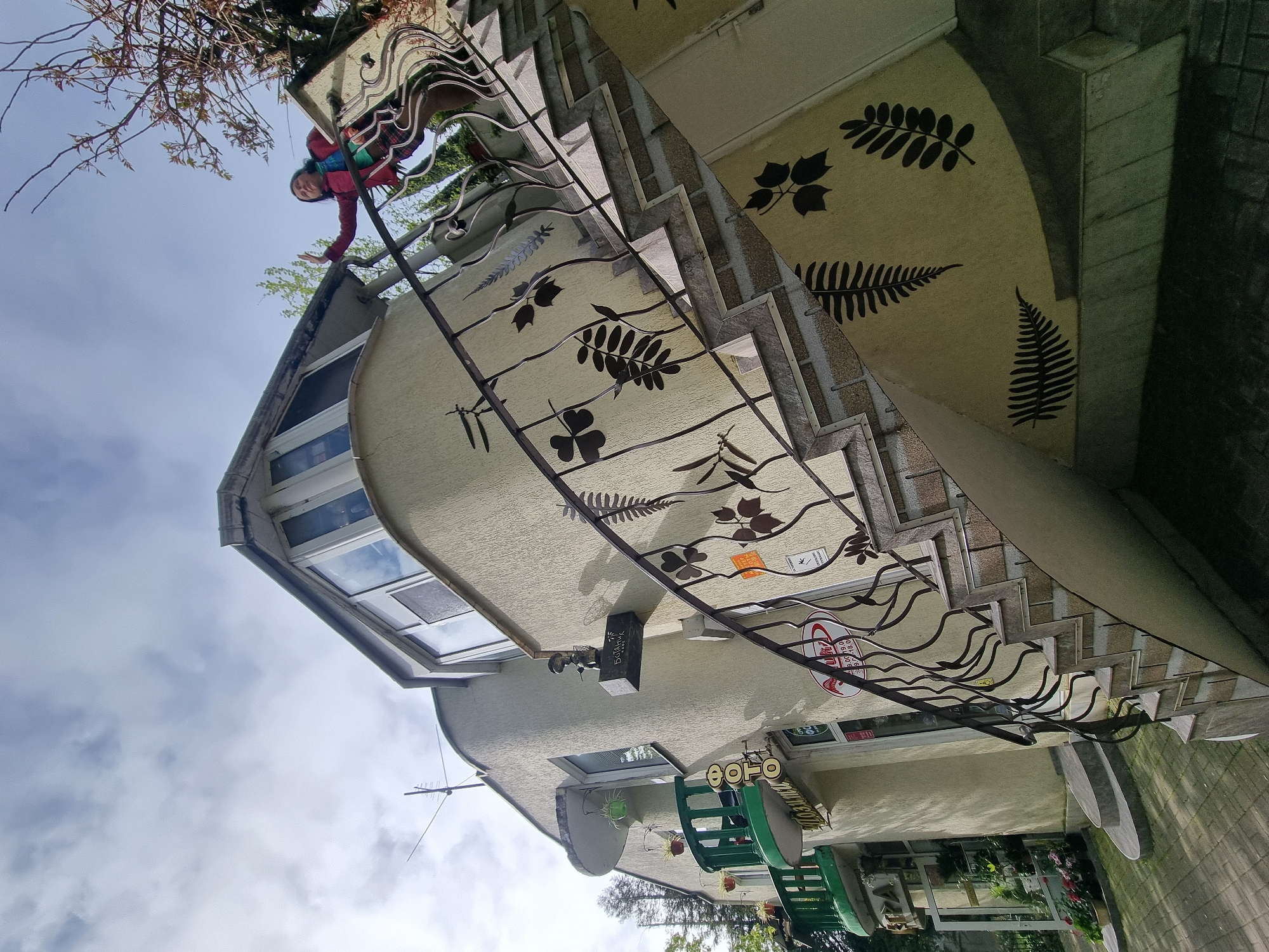 The image shows a multi-story building with unique architectural details, located in Sochi, Russia. The building has a light-colored facade adorned with black fern leaf patterns. There is a prominent staircase with an ornate metal railing featuring botanical designs. A person is visible near the top of the building, appearing to be reaching over the railing. The sky is partly cloudy. The lower floors have windows, signage, and decorative elements like hanging planters. There are chairs visible as well. The building’s style is distinct.