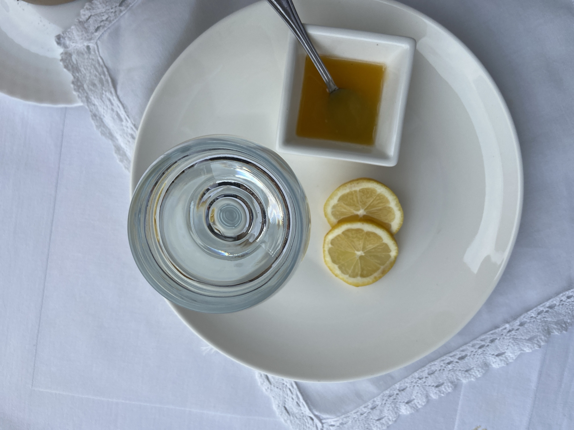 A still life taken from above shows a white plate on a white linen tablecloth, likely indoors. On the plate is a water glass filled with clear water, a small square bowl of honey with a spoon in it, and two lemon slices. Part of another white plate is visible in the upper left corner of the frame. The tablecloth is decorated with white lace. The location, Budva, Montenegro, is not explicitly suggested by the elements in the picture.