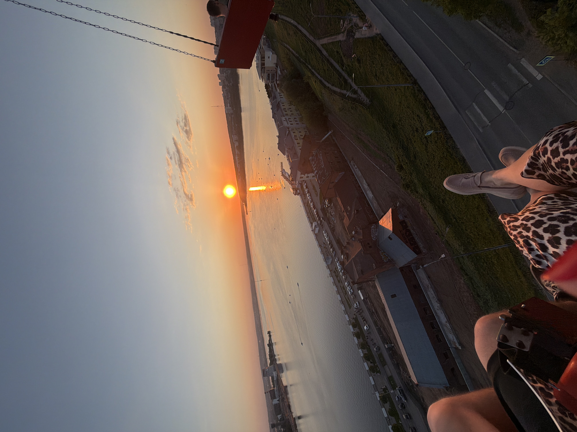 View from a rotating swing ride in Izhevsk, Russia, possibly at the Tsentralnaya Ploshchad (Central Square) during sunset. The swing car is visible in the lower right and upper left corners. The rider's legs, feet, and shoes are in the foreground. In the mid-ground is the Votkinsk Pond, reflecting the setting sun. The city buildings, road, and green space are parallel to the pond. The sky is predominantly orange and blue.