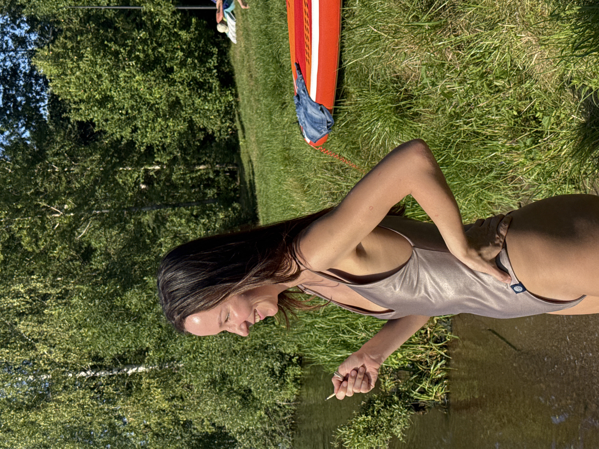 A woman in a metallic one-piece swimsuit stands by the edge of a body of water, holding a small object in her right hand and with her left hand on her hip. She is looking to her left and smiling. Behind her, there are trees and a grassy slope. On the grass, an orange stand-up paddleboard with a blue shirt on it is visible, along with two other people in the background. The scene appears to be outdoors on a sunny day. This image was taken in Moscow, Russia.