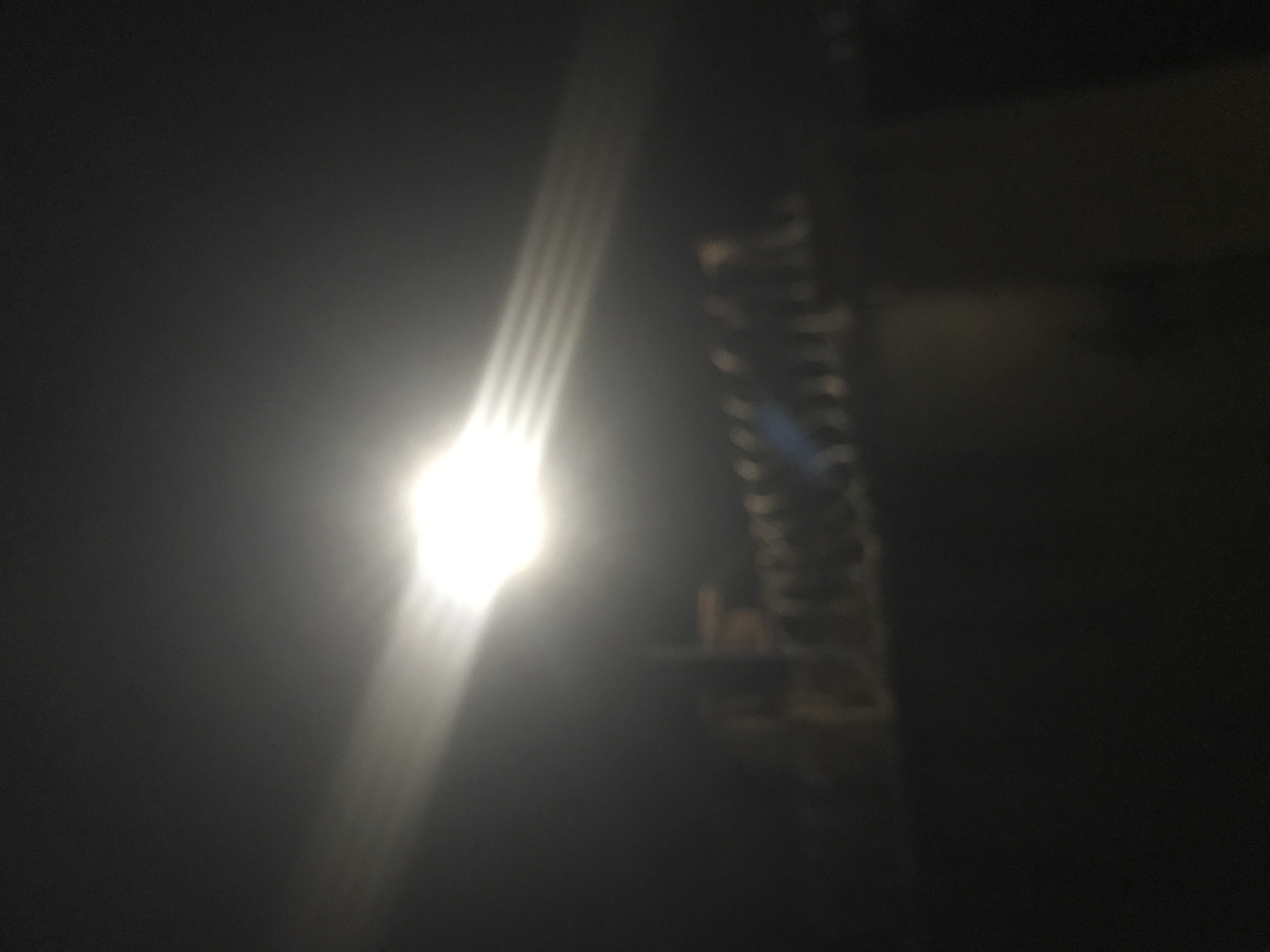 The image depicts a blurry nighttime scene. A bright light source, possibly a street lamp or searchlight, is visible in the upper left quadrant, casting strong horizontal beams of light across the frame. Below the light, a dark, out-of-focus structure is visible, resembling a railing or a fence with indistinct figures or posts along it. The overall environment appears to be an outdoor urban setting at night, with a lack of sharp detail due to the low light and blur. No specific landmarks or identifiable features of Rafin Par, Nigeria, are discernible.