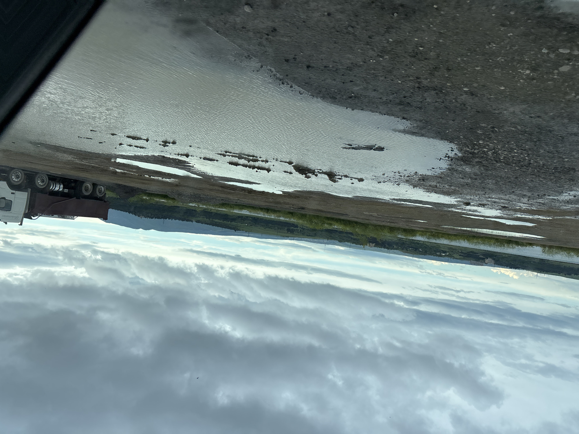The image shows a landscape with the foreground dominated by a muddy, waterlogged area near a road. A heavy truck is visible on the left. In the middle ground, there is a band of green vegetation along a body of water. Beyond that, a low mountain range is visible. The sky is filled with cloudy weather. The location appears to be outside the city of Tbilisi, Georgia, near the Tbilisi Sea.