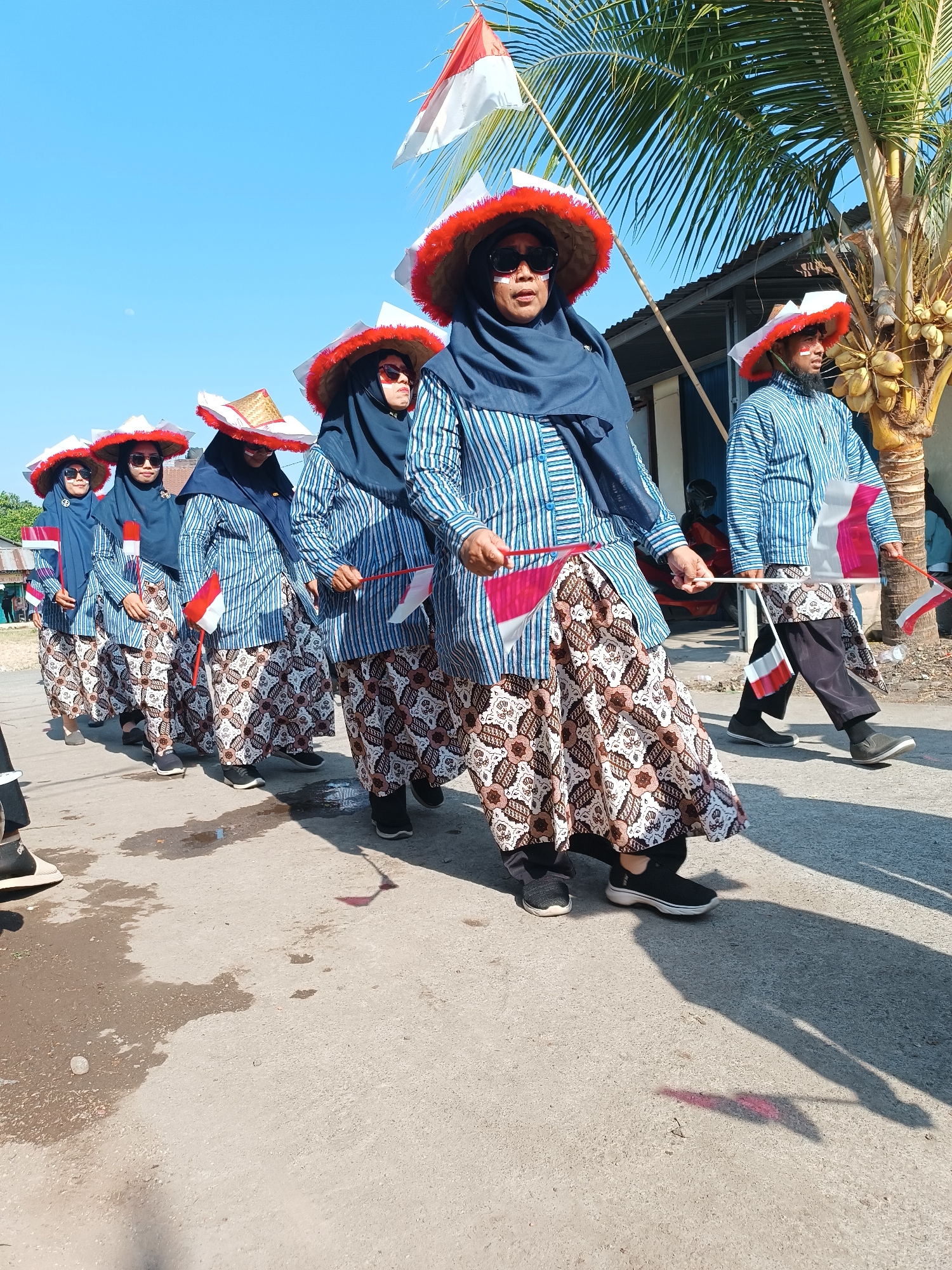 In Imogiri, Indonesia, a group of six individuals, predominantly women, are participating in a procession. They are dressed in traditional Indonesian attire, featuring blue and white striped shirts and patterned batik skirts. Each person wears a wide-brimmed hat adorned with red trim and a dark blue headscarf. Many are holding small Indonesian flags. The procession appears to be taking place on a paved road under a clear blue sky, with a palm tree and a building visible in the background. Some individuals have decorative white face paint. The scene suggests a festive or celebratory event, possibly related to a national holiday.
