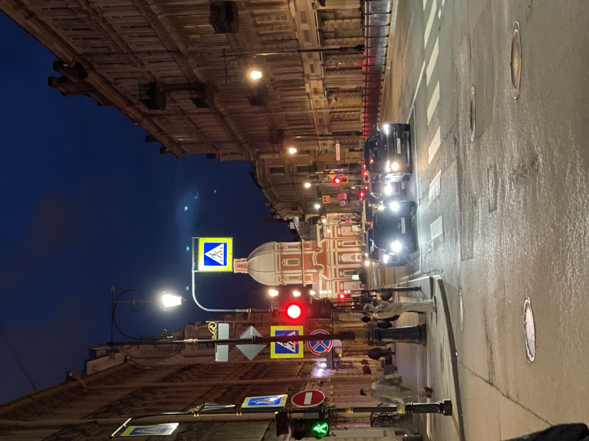 The image shows a street scene at night in Saint Petersburg, Russia. The street is lined with multi-story buildings with traditional architecture. A red and white domed building, likely a church or cathedral, is visible in the background. Cars are driving on the wet street, and pedestrian crossings are marked with white lines. Traffic lights are visible, with a red light illuminated. Several street lamps are lit, casting a warm glow on the surroundings. Pedestrians are walking on the sidewalk. There are also traffic signs and street furniture present. The sky is dark blue, indicating it is nighttime. The overall atmosphere is that of an urban environment in the evening.