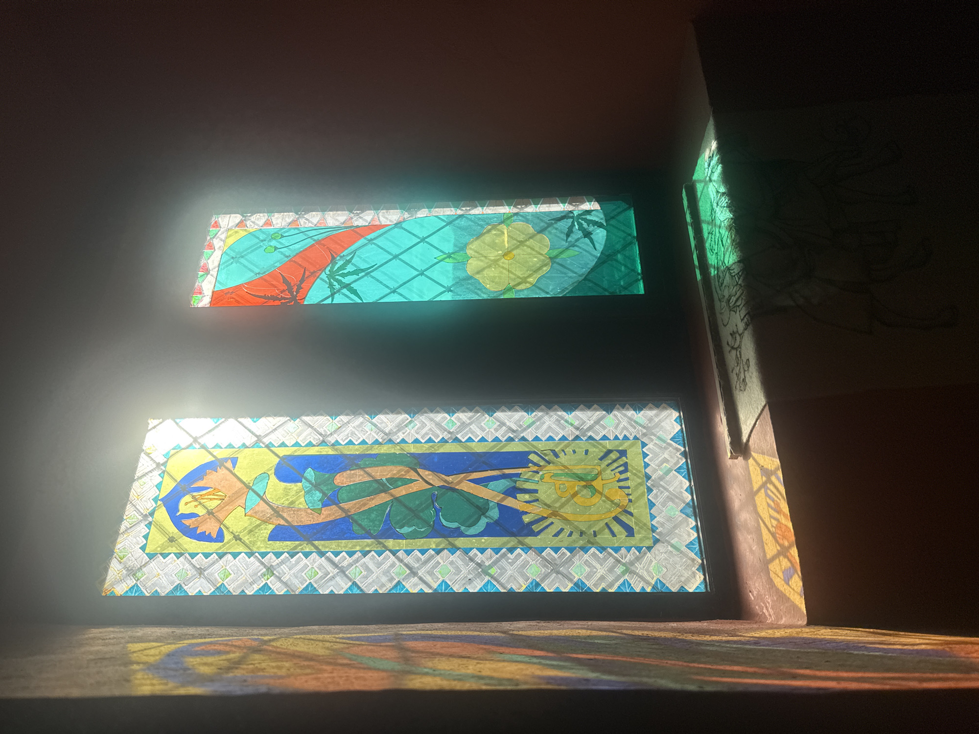 Two rectangular stained-glass windows are seen in an enclosed space, likely a room, casting colorful patterns of light onto the floor. The top window features a turquoise background with a red curved shape and a yellow flower. The bottom window has a yellow background with blue and green abstract shapes, possibly depicting stylized plants or figures. The windows are framed with a geometric pattern. Sunlight shines through the windows, projecting hues of orange, yellow, and blue onto the floor and walls. An adjacent wall has faint drawings sketched onto it. The location is Chynadiiovo, Ukraine.