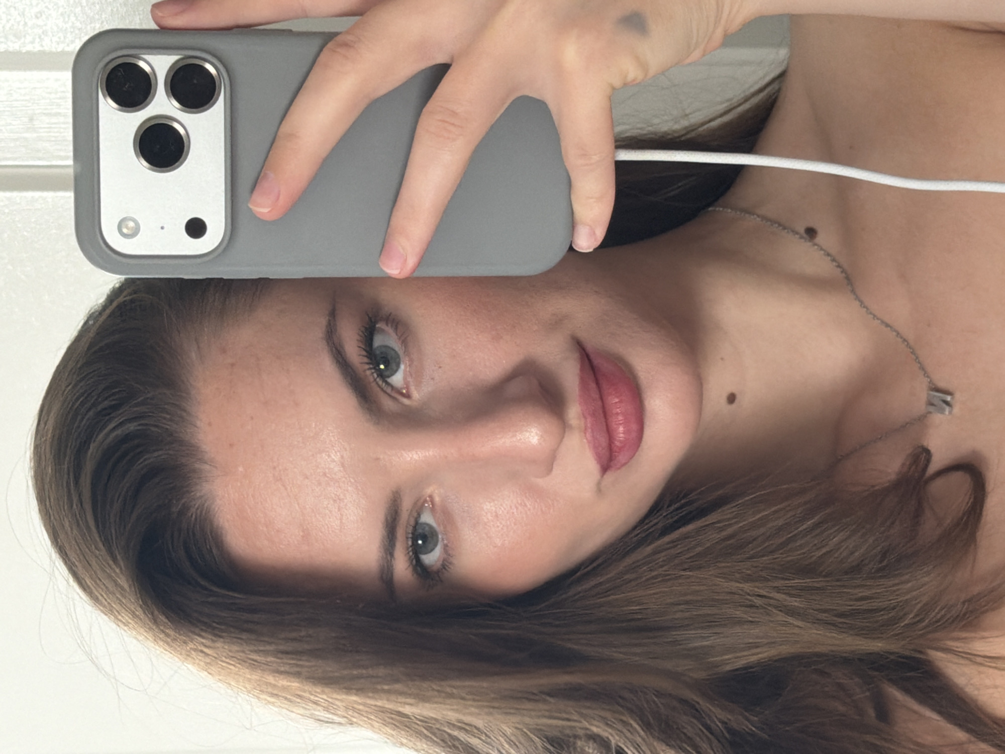 A young woman with brown hair and blue eyes is taking a selfie with her iPhone. She is wearing a grey phone case and a silver necklace with a charm. She has a mole on her neck and a tattoo on her arm. She is indoors, in front of a white door. The lighting suggests it is daytime.