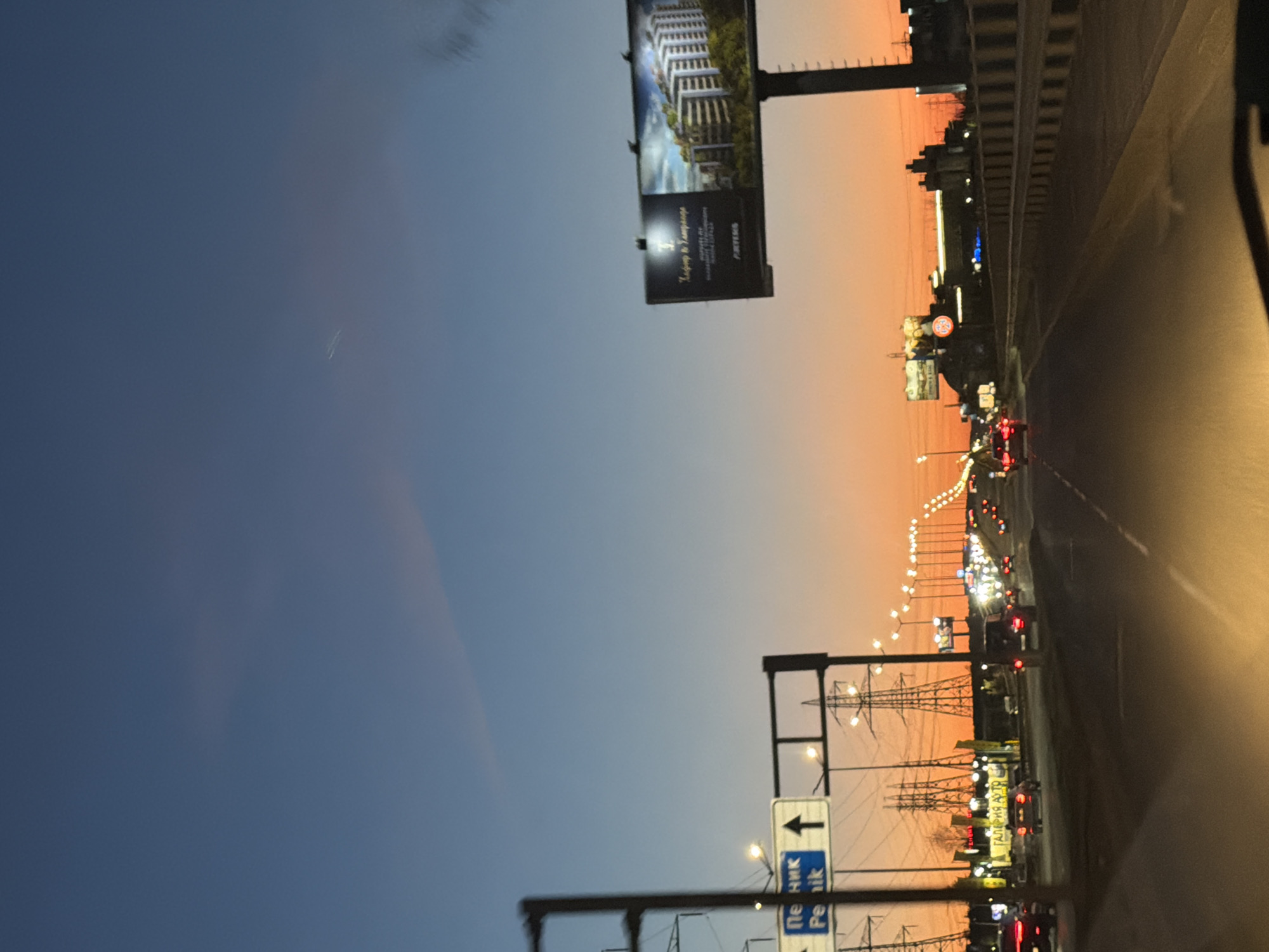 The image shows a road with traffic at dusk or dawn, likely in Sofia, Bulgaria, based on the Cyrillic script on the road signs and billboards. The sky transitions from a dark blue to orange. On the left side of the road, there are billboards and overhead power lines. Cars with their headlights and taillights on are driving on the road. The view is likely from inside a vehicle, with a portion of the vehicle's interior visible on the right.
