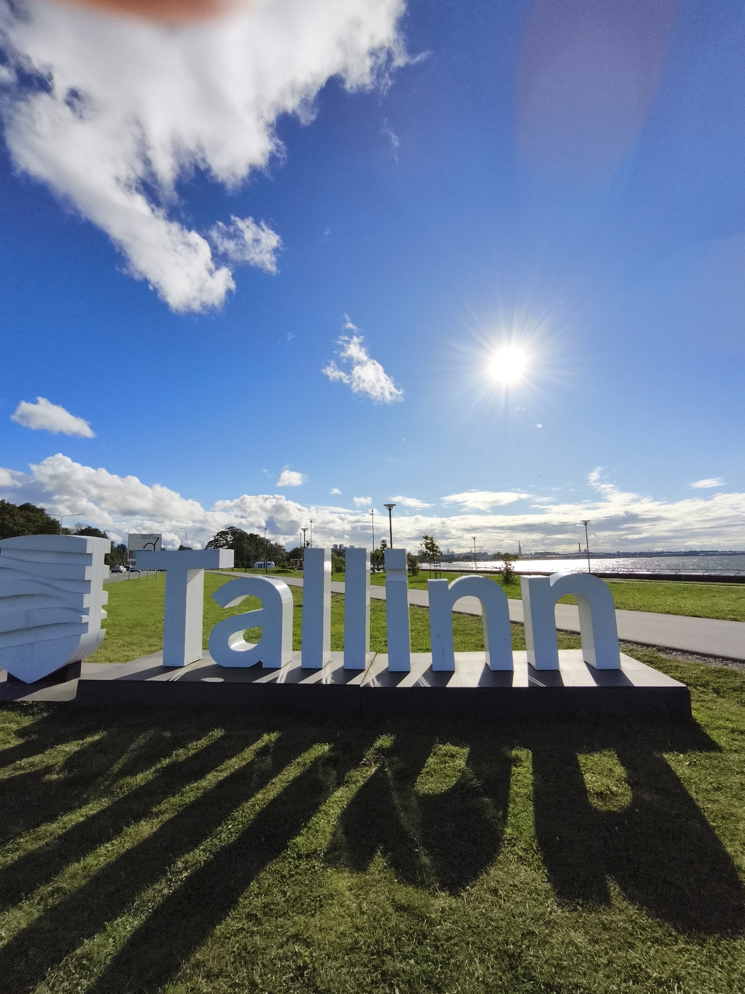 This is a wide-angle, low-angle outdoor photograph of a large, white, three-dimensional sculpture spelling out "Tallinn" in front of a body of water and a bright blue sky. The sun is prominently visible in the upper right quadrant of the sky, creating lens flare and casting long shadows of the sculpture onto the green grass in the foreground. The sculpture is positioned on a dark platform, which is set on a lawn. Behind the sculpture, a paved walkway runs parallel to the water. In the distance, across the water, a city skyline with buildings and structures is visible. There are streetlights and trees lining the waterfront area. The sky is partly cloudy with scattered white clouds. The image appears to be taken on a sunny day. The "Tallinn" sculpture is a recognizable landmark in Tallinn, Estonia, often found near the waterfront.