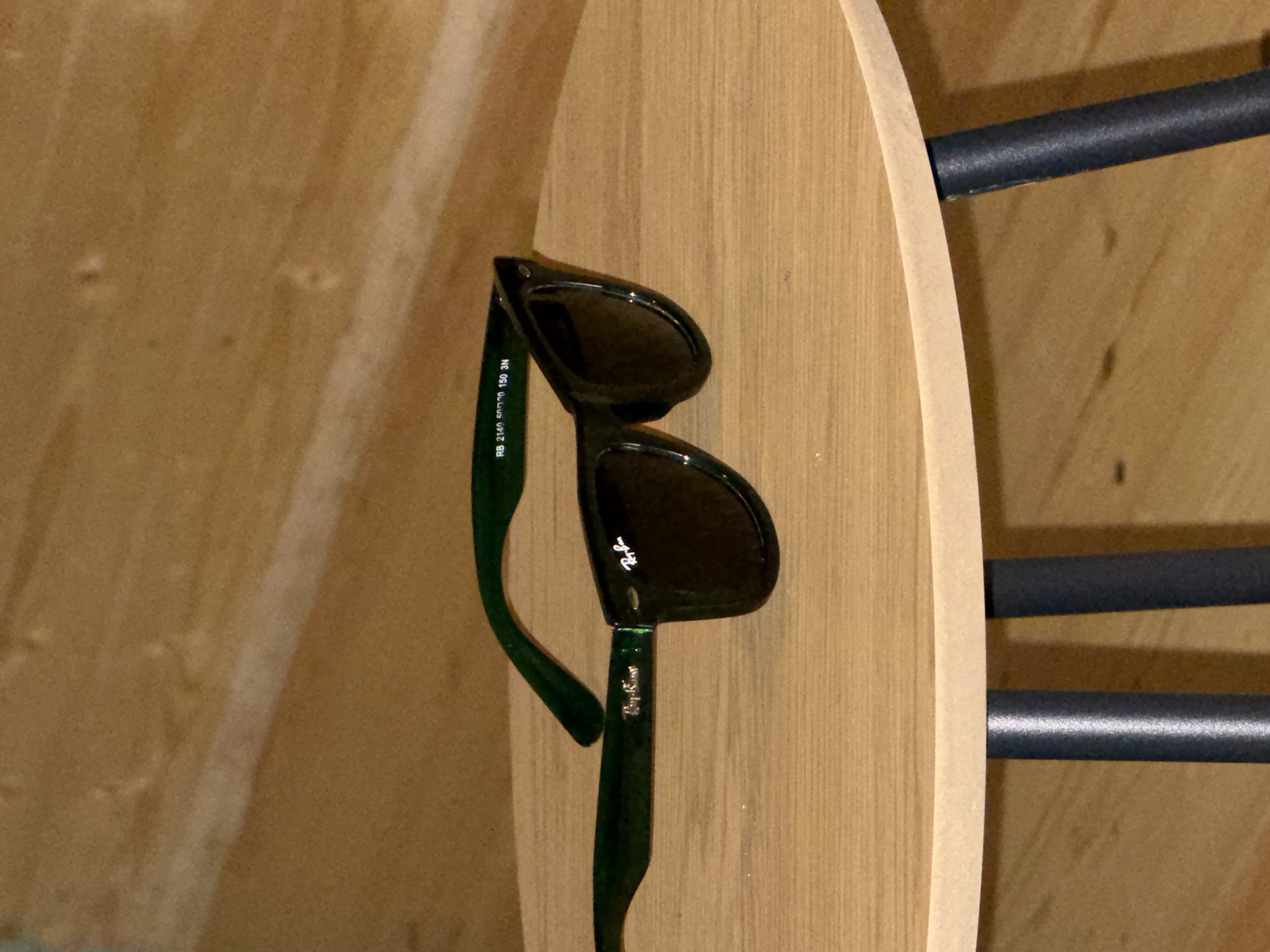 A pair of dark green Ray-Ban Wayfarer sunglasses rests on a light brown wooden tabletop. The sunglasses are positioned to the left of the frame, with the lenses facing the viewer and the arms folded. The left arm of the sunglasses is visible, extending towards the right side of the image, while the right arm is mostly obscured. The tabletop appears to be part of a small, round table, supported by dark-colored legs that are partially visible at the bottom of the frame. The background consists of a textured, light brown wooden surface, likely a wall or partition. The lighting is dim, creating shadows and highlighting the texture of the wood. No people or other objects are present in the scene. The location is Unknown, Unknown.