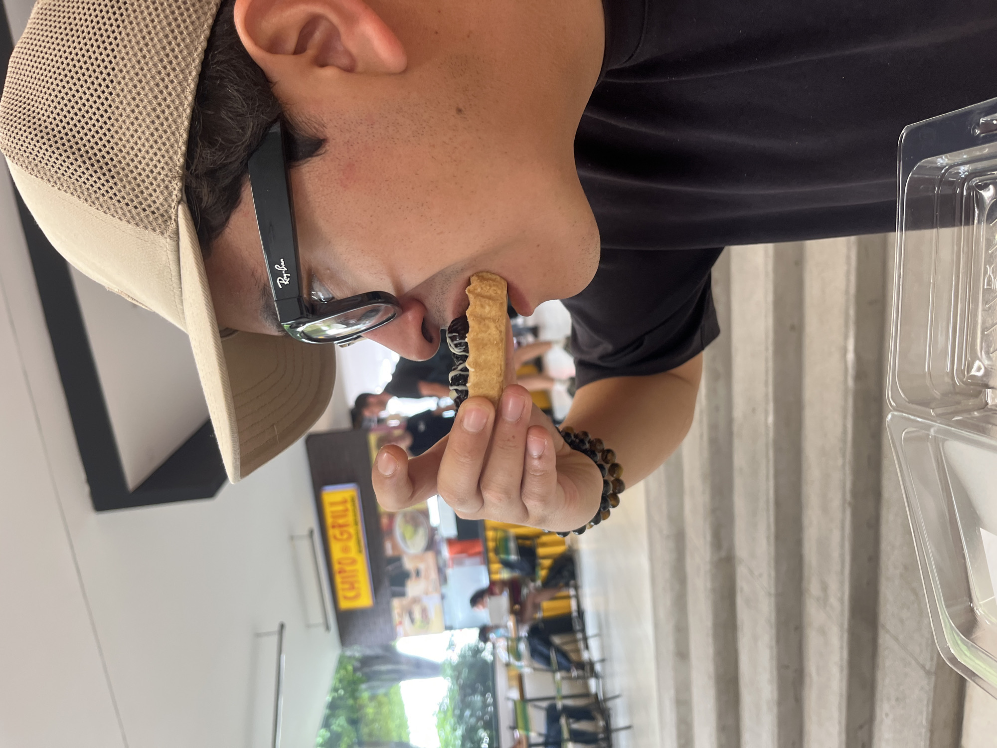 An image, captured in San Andrés Cholula, Mexico, features a person in profile within a casual dining or food court environment. The subject, viewed from the right, wears a beige mesh baseball cap, black-framed Ray-Ban sunglasses, and a black t-shirt. They have dark hair and visible facial stubble.
The person is actively holding a golden-brown, ridged pastry, resembling a churro, to their mouth, seemingly in the process of eating it. The pastry is topped with dark, spherical additions, possibly chocolate pearls or boba. On their right wrist, a beaded bracelet composed of brown and black stones is visible.
The background is softly blurred, revealing a public dining area. A bright yellow sign with the red text "CHITO GRILL" is visible to the left, indicating a food vendor. Several other individuals are seated at tables in the distance, enjoying meals. Architectural features include light-colored concrete steps or slatted walls, and a glimpse of greenery suggests an open or brightly lit setting. A clear plastic food container is partially visible on a surface to the right. The scene depicts a relaxed moment during a daytime meal.
