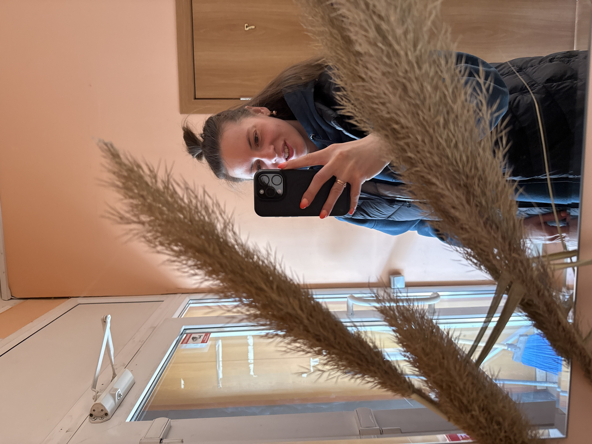 A woman takes a selfie in a mirror. The mirror reflects a door with a hydraulic door closer and a sign. The woman is wearing a dark jacket and has orange nail polish. There are tall, dried flower stalks in front of the mirror. A blue broom is visible through the door glass. Given the architectural details and the style of the door hardware, the location could potentially be within the city of Kazan, Russia.
