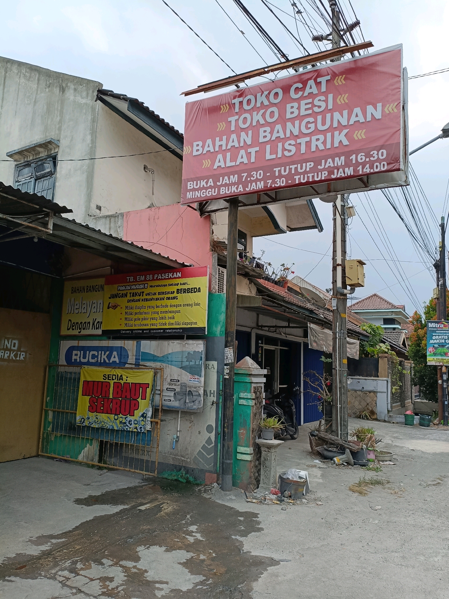 This is a medium-distance, eye-level, outdoor photograph of a street scene in Maguwoharjo, Indonesia. A prominent red and white banner hangs over the street, advertising "TOKO CAT TOKO BESI BAHAN BANGUNAN ALAT LISTRIK" (Paint Store, Iron Store, Building Materials, Electrical Tools). The banner also provides opening hours: Monday to Saturday 7:30 AM to 4:30 PM, and Sunday 7:30 AM to 2:00 PM.
Below the large banner, another sign in yellow and red reads "TB. EM 88 PASEKAN" and details several points, including "JANGAN TAKUT UNTUK BERSIKAP BERBEDA DENGAN KEBIJAKAN ORANG" (Don't be afraid to be different from others' policies) and lists several principles for customer service.
To the left, a metal gate with green paint is visible, with a yellow sign hanging on it that says "SEDIA: MUR BAUT SEKRUP" (Available: Screws, Bolts, Nuts). A portion of a building with a corrugated metal roof is to the left of the gate.
Further down the street, a blue storefront is visible, and a motorcycle is parked in front of it. Utility poles with numerous wires are scattered throughout the scene, indicating an urban or semi-urban environment. The ground is paved with concrete and shows signs of water or oil stains. There is also some debris on the ground, including buckets and plant pots. In the background, residential buildings with tiled roofs are visible. A small sign for "Mitra" is visible on the right side of the image. The sky is overcast. No people are clearly visible in the frame.
