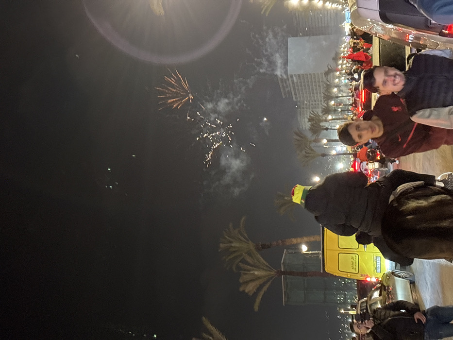 Night scene with fireworks in the sky. Palm trees line the street. People are gathered, some standing by cars. A yellow van is visible. Individuals are looking towards the fireworks. Buildings can be seen in the background, with lights illuminating the scene. The overall atmosphere is festive and celebratory.