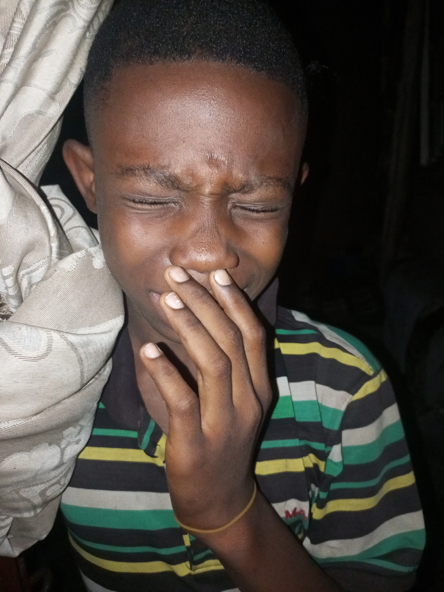 A close-up, medium shot, in a dark room, shows a young Black boy with his eyes closed and his hand covering his mouth, possibly crying or distressed. He is wearing a striped shirt with black, white, green, and yellow bands. To the left of the boy, a light-colored cloth with a pattern is draped, obscuring part of his face and head. The background is dark and indistinct, suggesting an indoor environment.