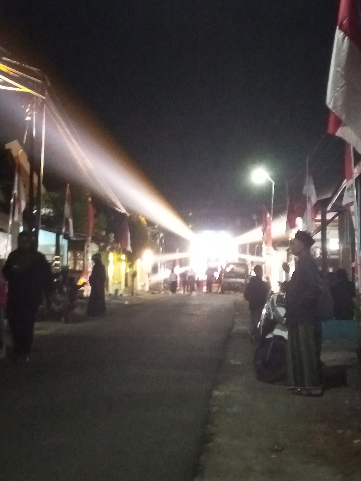 The image depicts a nighttime street scene in Turen, Indonesia, possibly during a local event or celebration. The lighting suggests a festive atmosphere, with bright stage lights illuminating a portion of the street and a prominent street lamp casting light on the scene. Several people are present, some standing and observing, while others appear to be walking or gathered. Flags, possibly Indonesian flags, are visible, suggesting a national or local observance. The presence of a vehicle in the background, along with the general layout of the street and buildings, indicates an urban or semi-urban environment. The overall impression is one of a community gathering or public event taking place after dark.