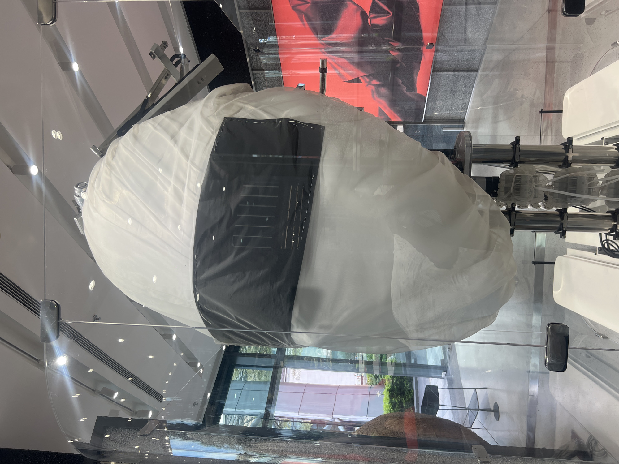 The image shows a large, oblong object resembling a wrapped rice ball or onigiri, encased in a transparent glass display case. The object is white and covered with a wide, black band in the middle. Within the black band are linear indentations or markings. The display case reflects the surrounding environment, including a view of a building facade with window panels, possibly exterior scenery with greenery, and interior elements such as lights and architectural details. The location is likely Tianhe District, China. The scene appears to be an exhibition or display within a public space like a museum or gallery. There are no people visible in the immediate frame. The focus is clearly on the displayed object itself.