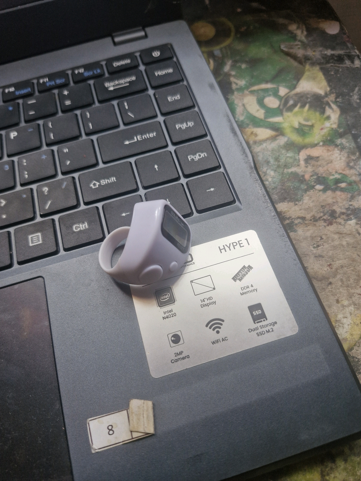 The image shows a close-up, eye-level view of a gray laptop keyboard and a digital counter ring placed on the keyboard. The laptop's surface is worn and appears to be on a textured, patterned surface. The digital counter ring is white and has a small screen and buttons. A sticker is present on the laptop, detailing specifications such as "HYPED 1", "14" HD Display", "DDR4 Memory", "SSD", "WIFI AC", "2MP Camera", and "Intel N4020". A separate sticker with the number "8" is adhered to the lower left of the laptop. The scene is illuminated by overhead light, creating subtle shadows. The context provided by the prompt indicates the location is Rantau, Indonesia, but there are no visual cues within the image to confirm this specific location.