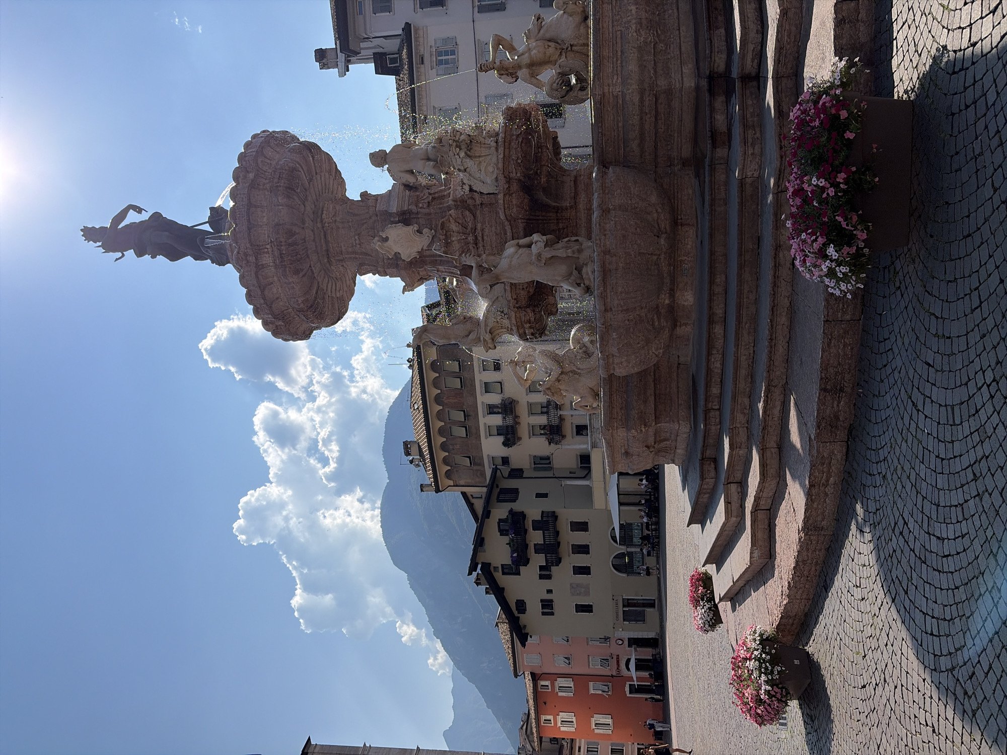 The image depicts a daytime scene in Trento, Italy, centered around a large, ornate fountain. The fountain features multiple tiers and intricate sculptures of figures and mythological creatures, with water spraying from various points. The sun shines brightly, casting shadows and highlighting the detail of the stone architecture.
In the background, buildings with multiple stories and balconies line a cobblestone street. Some of these buildings appear to be shops or cafes, with awnings and outdoor seating visible. Mountains rise in the distance, partially obscured by clouds, suggesting a pleasant weather day with some cumulus cloud formations.
Potted flowers in shades of pink and white add color to the scene, placed near the base of the fountain and along the cobblestone walkway. The overall atmosphere is one of a lively European town square on a sunny day. While individual people are visible, they are small in the frame and do not appear to be the primary focus. There is no discernible text in the image.