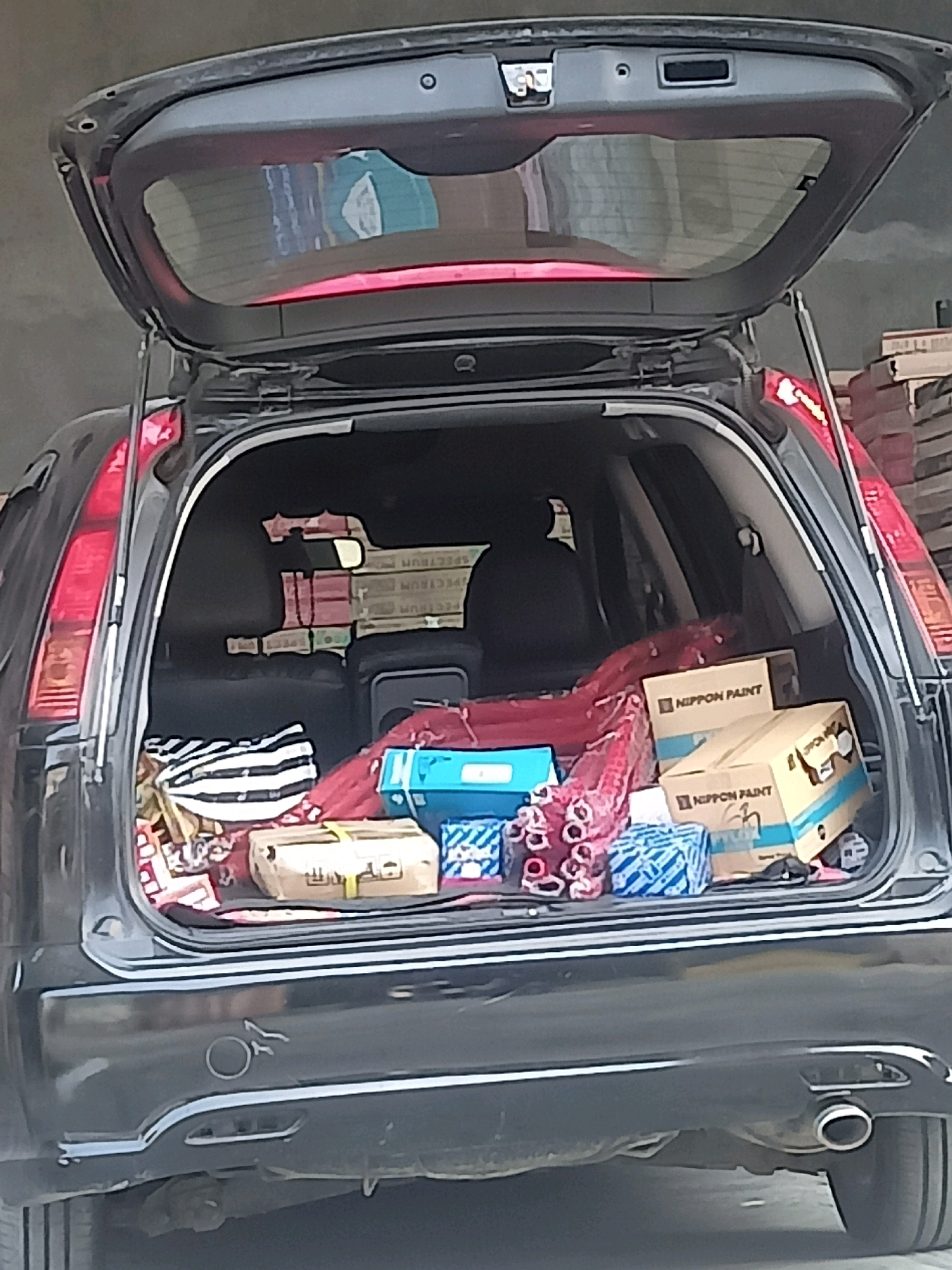 The image shows the open trunk of a dark-colored SUV parked in what appears to be a residential or commercial area in Karangtalun, Indonesia. The trunk is filled with various items, including several cardboard boxes, one of which is labeled "NIPPON PAINT." There is also a black and white striped bag, a red fabric item, and other packages wrapped in blue and white paper. The background is blurred, but it suggests an outdoor setting with some buildings or structures visible. The overall scene appears to be a common activity of loading or unloading goods into a vehicle.